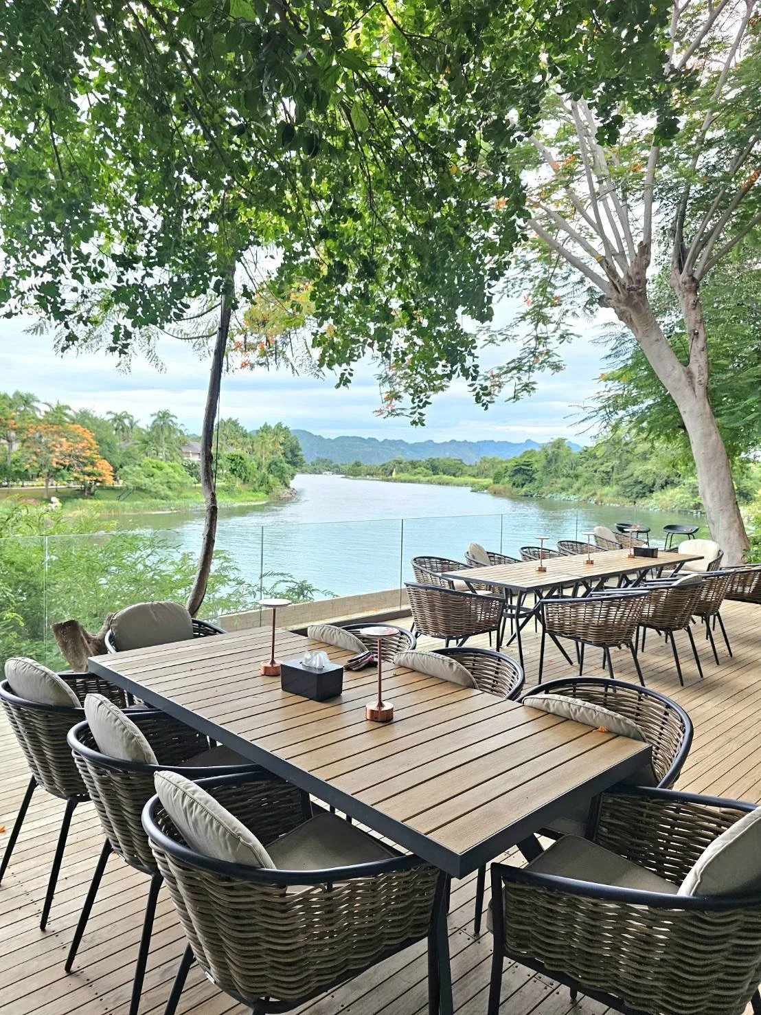 Outdoor balcony area with wooden tables and wicker chairs overlooking River Kwai bridge and River Kwai surrounded by green trees and distant mountains.