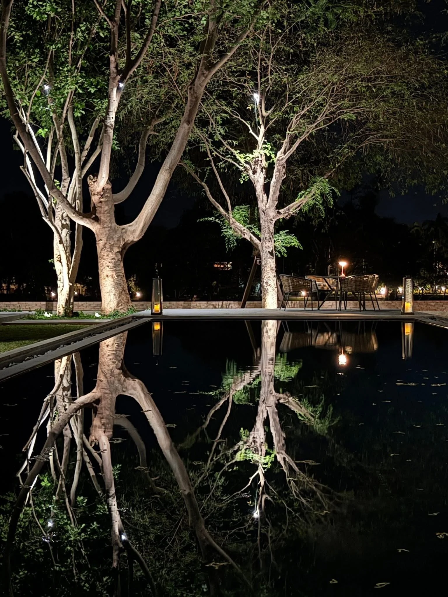 Night scene of trees with lights illuminating them and a reflection of the trees and lights in a body of swimming pool on riverside of River Kwai.