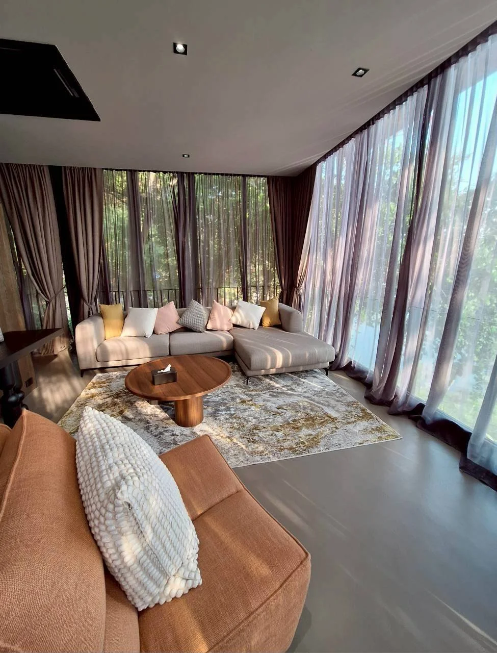 A modern Makham River View Suite living room with a beige sectional sofa, colorful throw pillows, a round wooden coffee table, and a burnt orange armchair with a textured white pillow, all set against large windows with sheer curtains letting in natu