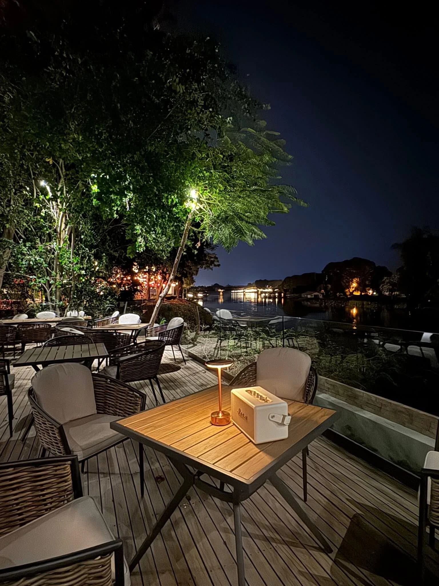 Outdoor patio seating area at night with wooden tables and chairs next to a body of water, surrounded by trees overlooking River Kwai bridge.