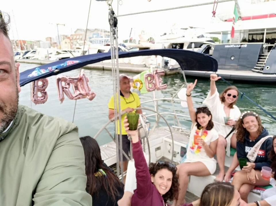 Gruppo di persone che festeggiano sulla barca, con decorazioni e brindisi, in un porto con barche e yacht.