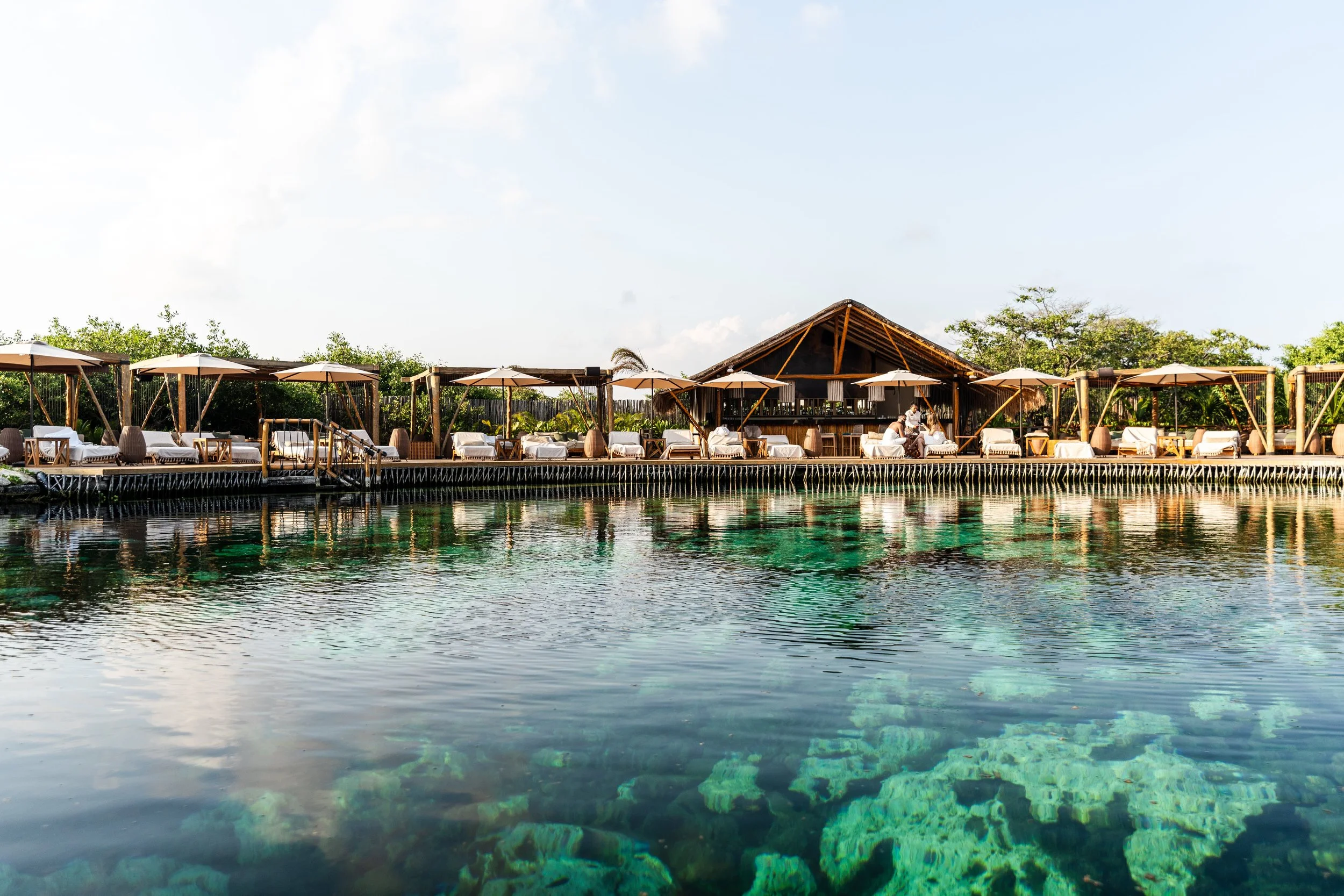 A scenic view of a resort with a deck overlooking clear water with visible coral and rocks. The deck has lounge chairs with white cushions, umbrellas, and a bar area, set against green trees and a blue sky.