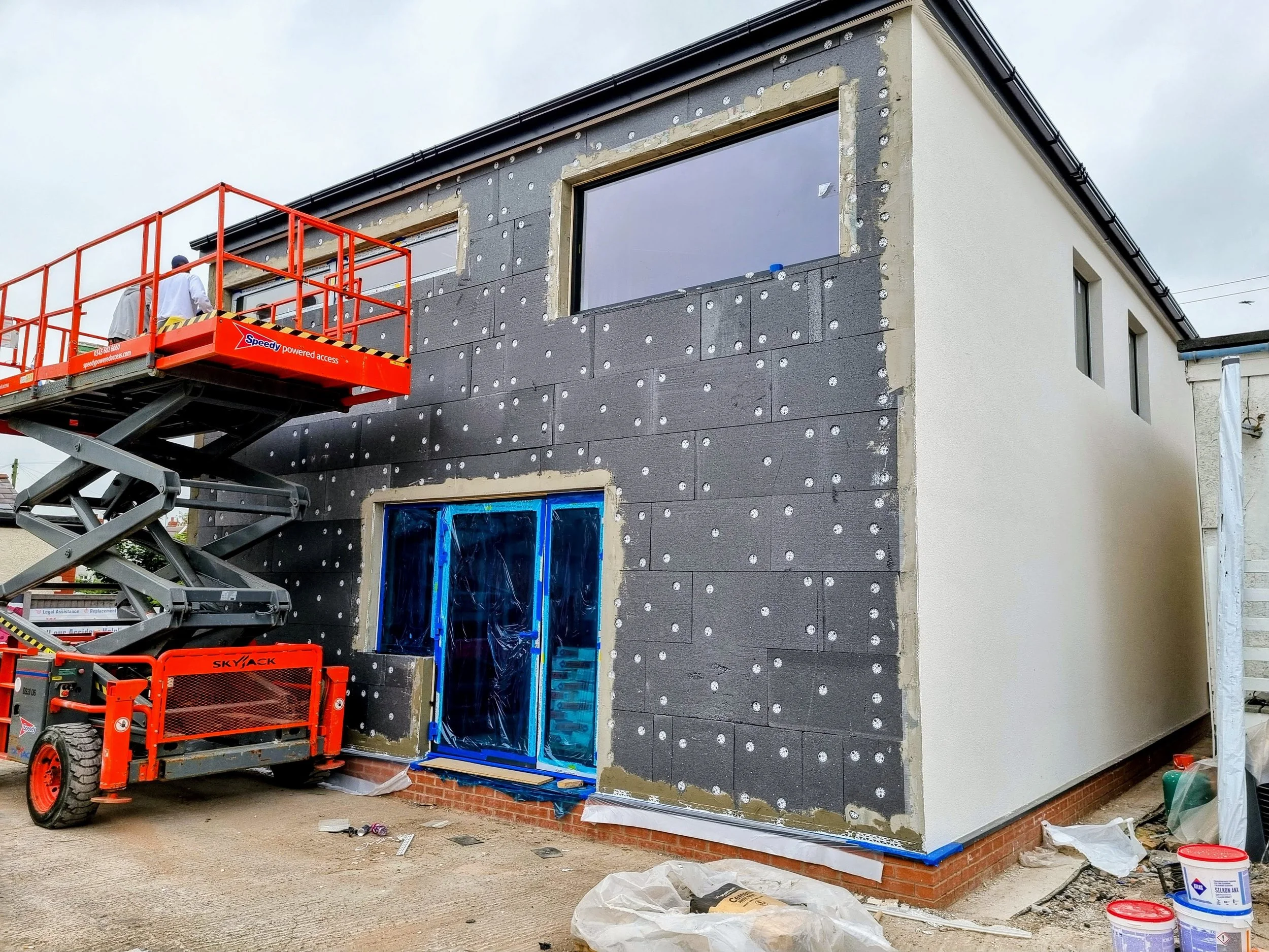 Construction workers are installing insulation and exterior finishing on a house, with a cherry picker lift positioned next to the building. render THIS