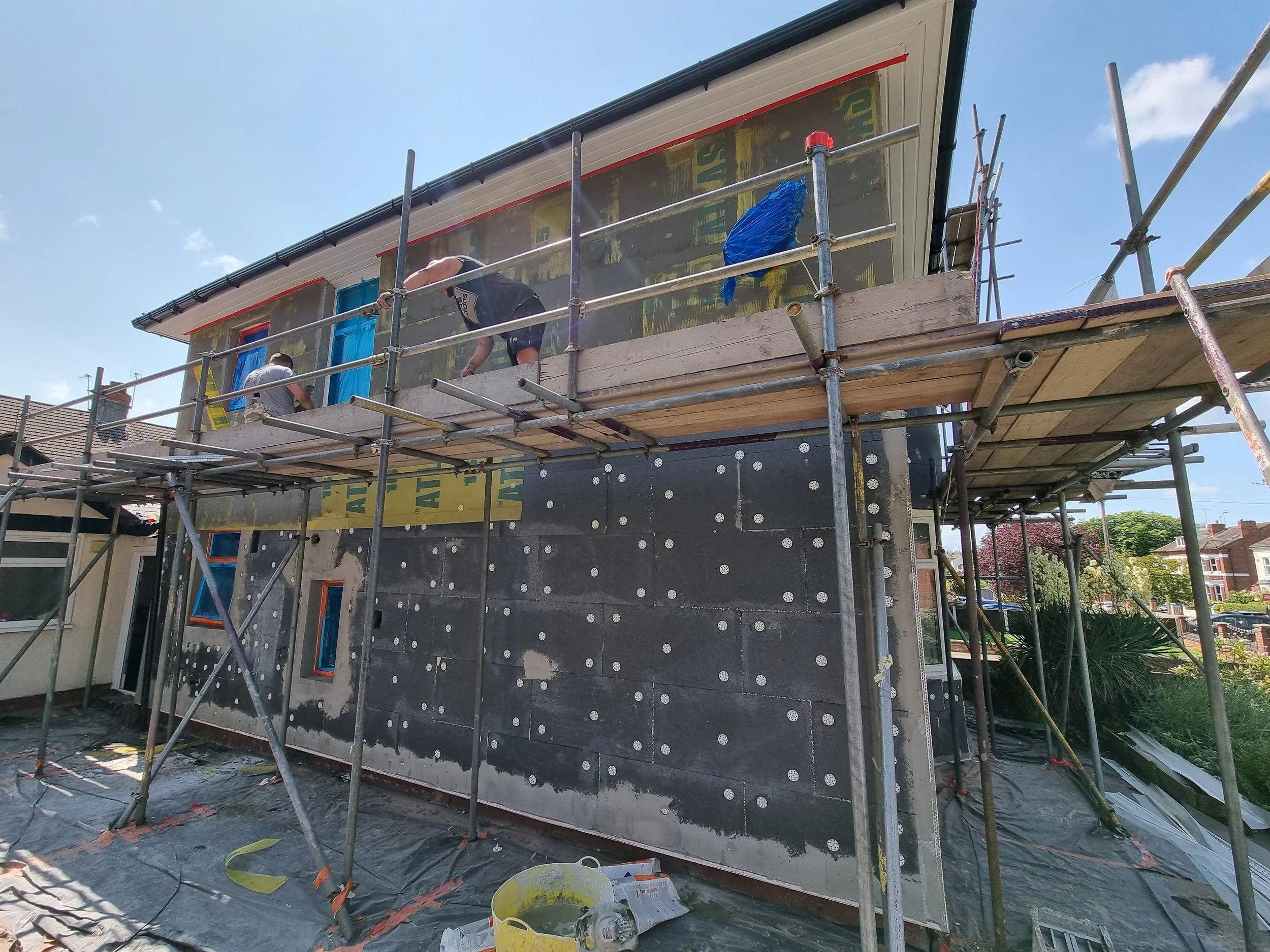 Construction workers on scaffolding installing exterior insulation and sheathing on a house wall during daytime.