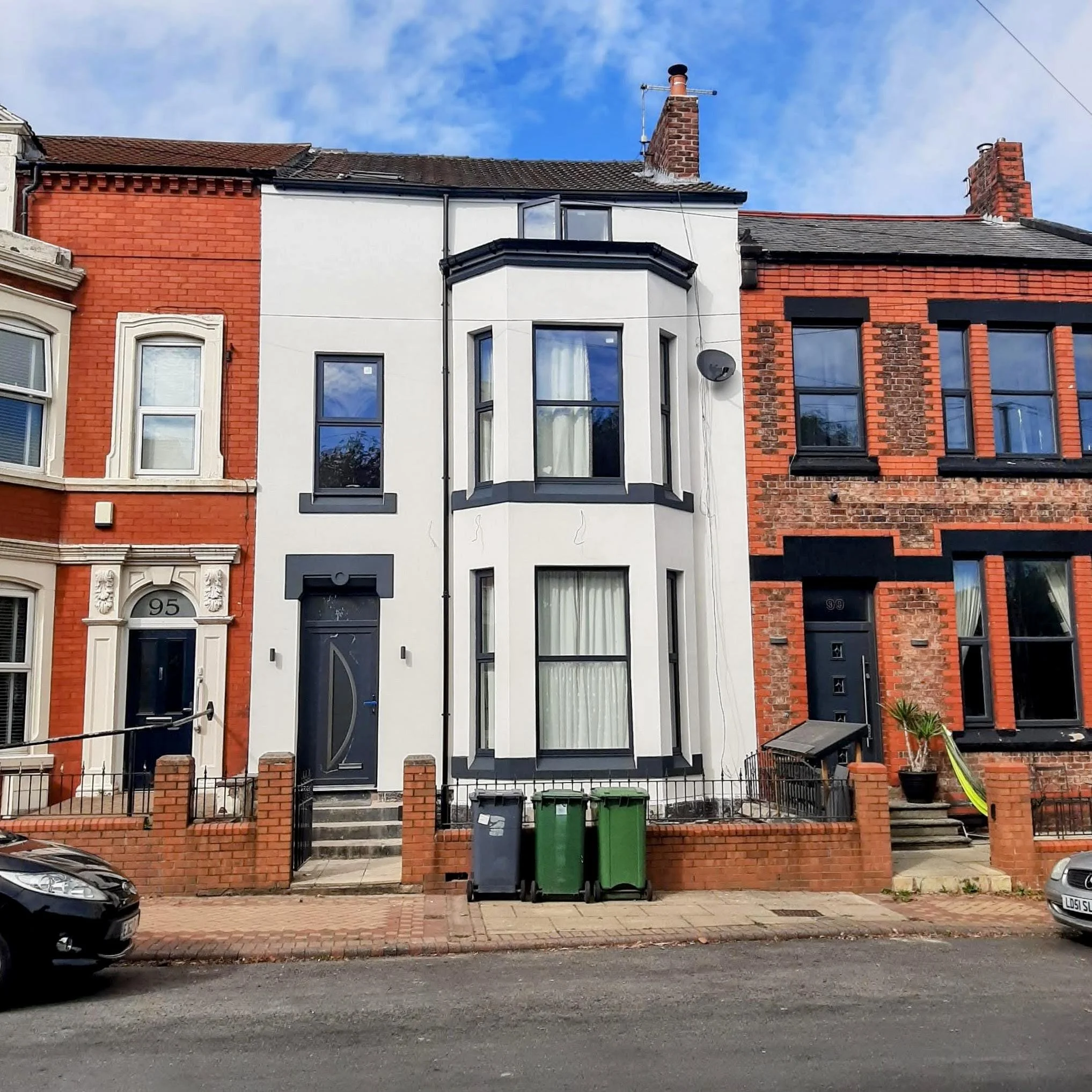 Three-story row house with a white facade, black window frames, and a curved black front door, situated between two red brick row houses, with trash bins in front. EWI, external wall insulation, brick slips, silicone render, K Rend