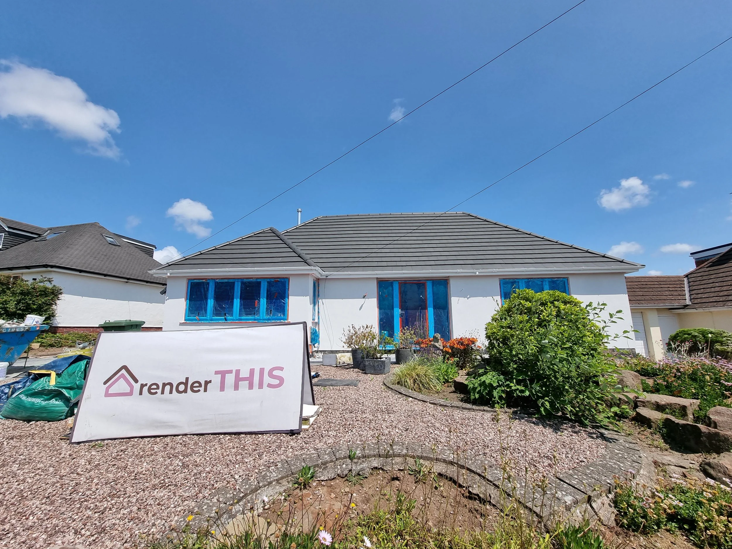A house with a newly renovated exterior, in silicone render, with windows and doors covered in blue painter's tape, indicating ongoing rendering or renovation work. A sign in the front yard reads 'render THIS'.
