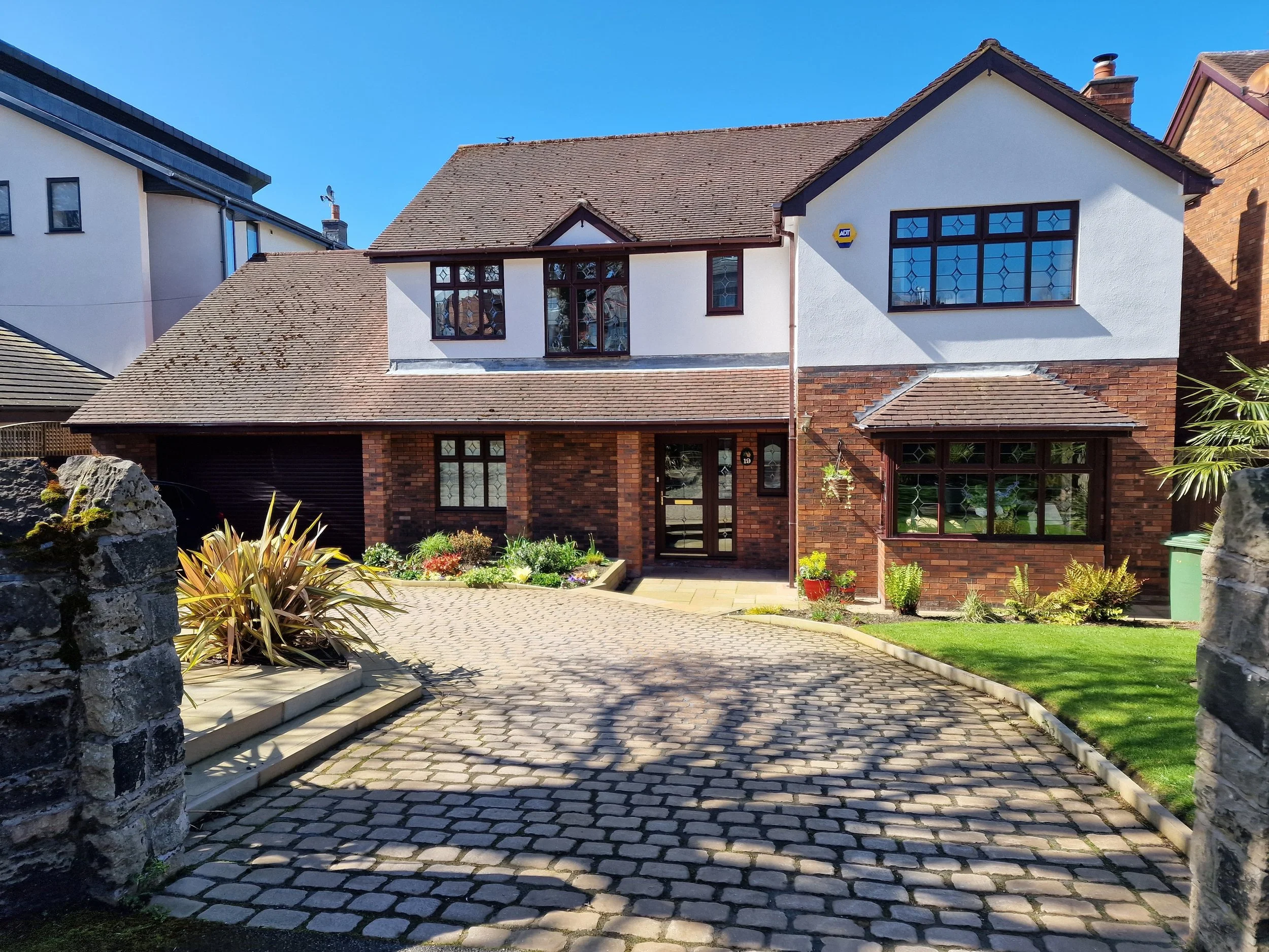 Front view of a two-story house with a brick and white exterior, cobblestone driveway, garden bed, and front lawn. Silicone render, K Rend, Rendering Liverpool, Wirral, renderers near me, house rendering, ATLAS render, thin coat render