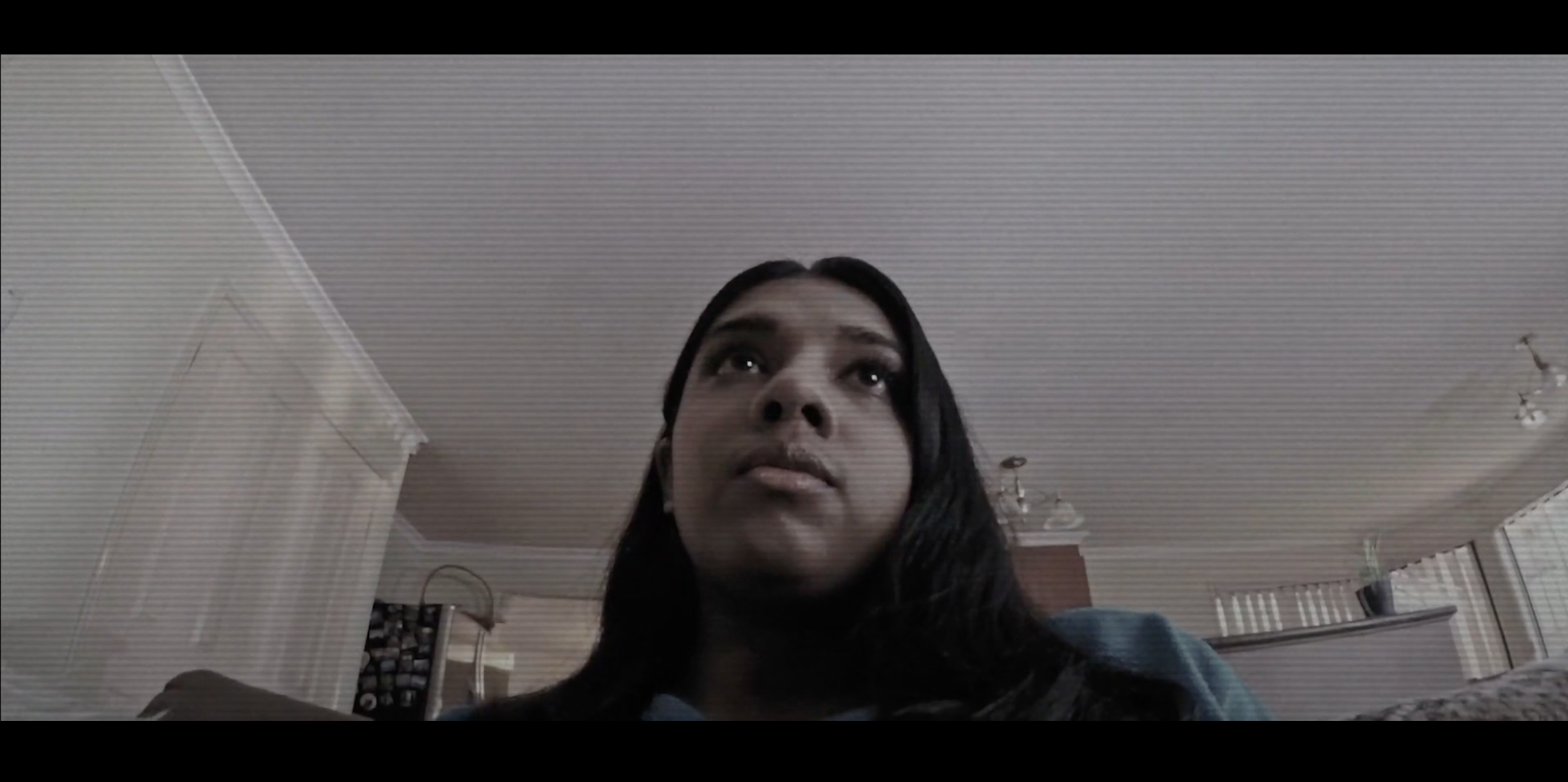 A woman with long dark hair looking upward in a room with white walls and ceiling, lit by ceiling lights, with some furniture and decor visible in the background.