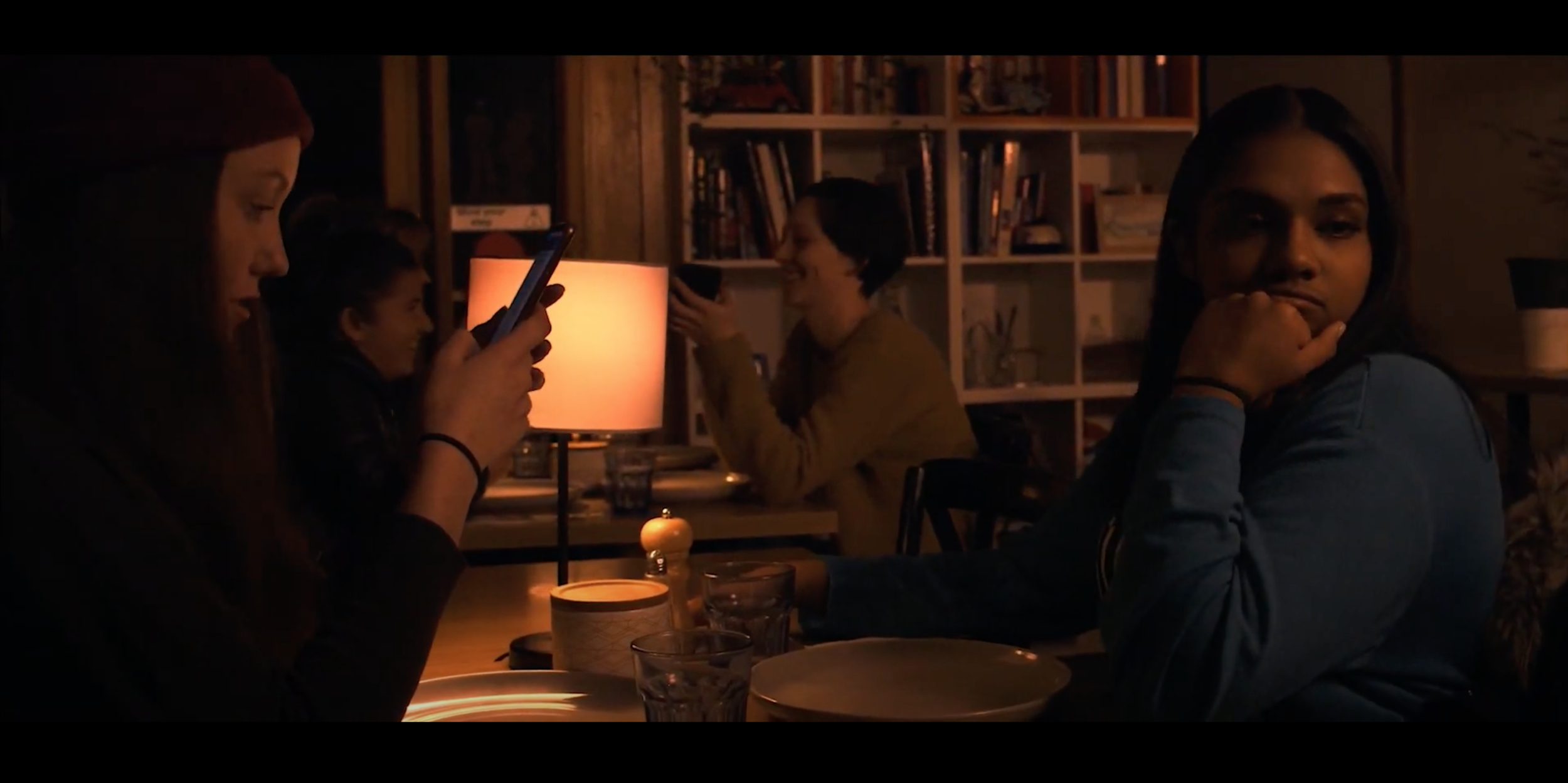 Four women sitting around a dining table in a cozy, dimly lit room, with bookshelves in the background. One woman is looking at her phone, another is smiling and holding a cup, and the others are resting their heads on their hands, engaged in different activities.