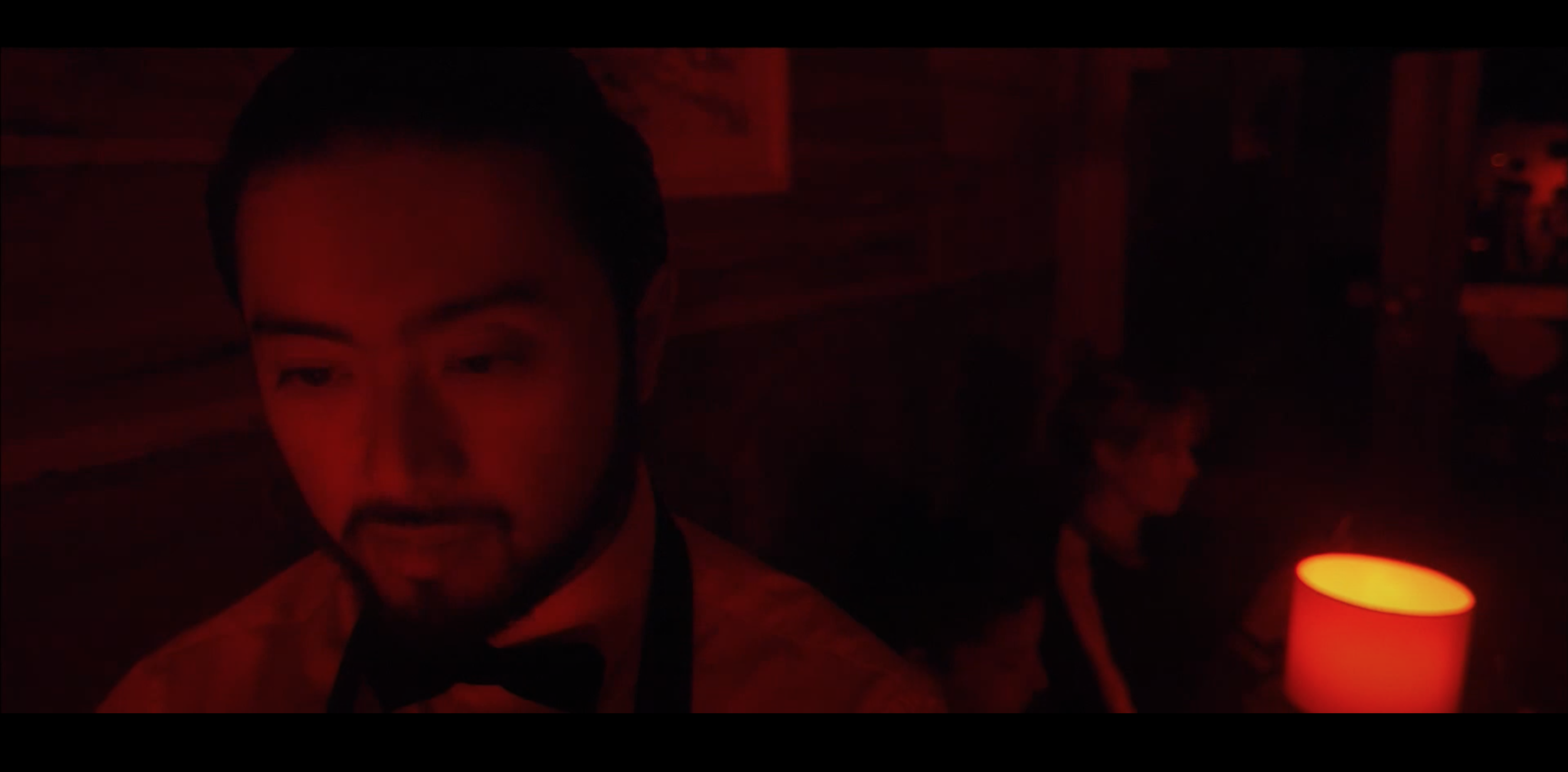 Man in formal attire with bow tie in dim, red-lit room, with a woman sitting in background next to a lit lamp.