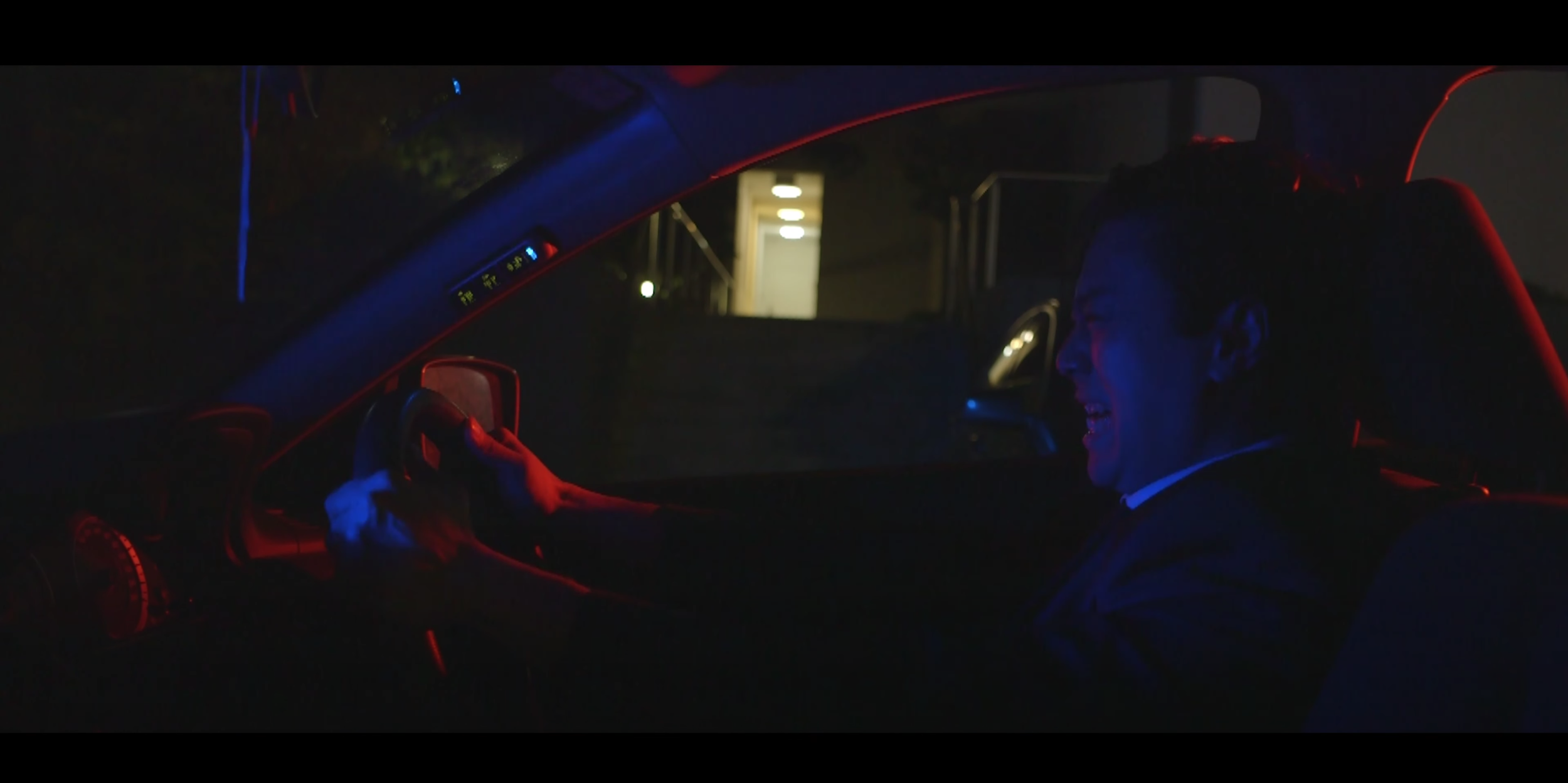 A person sitting in a car at night, smiling and holding the steering wheel, with red and blue interior lighting