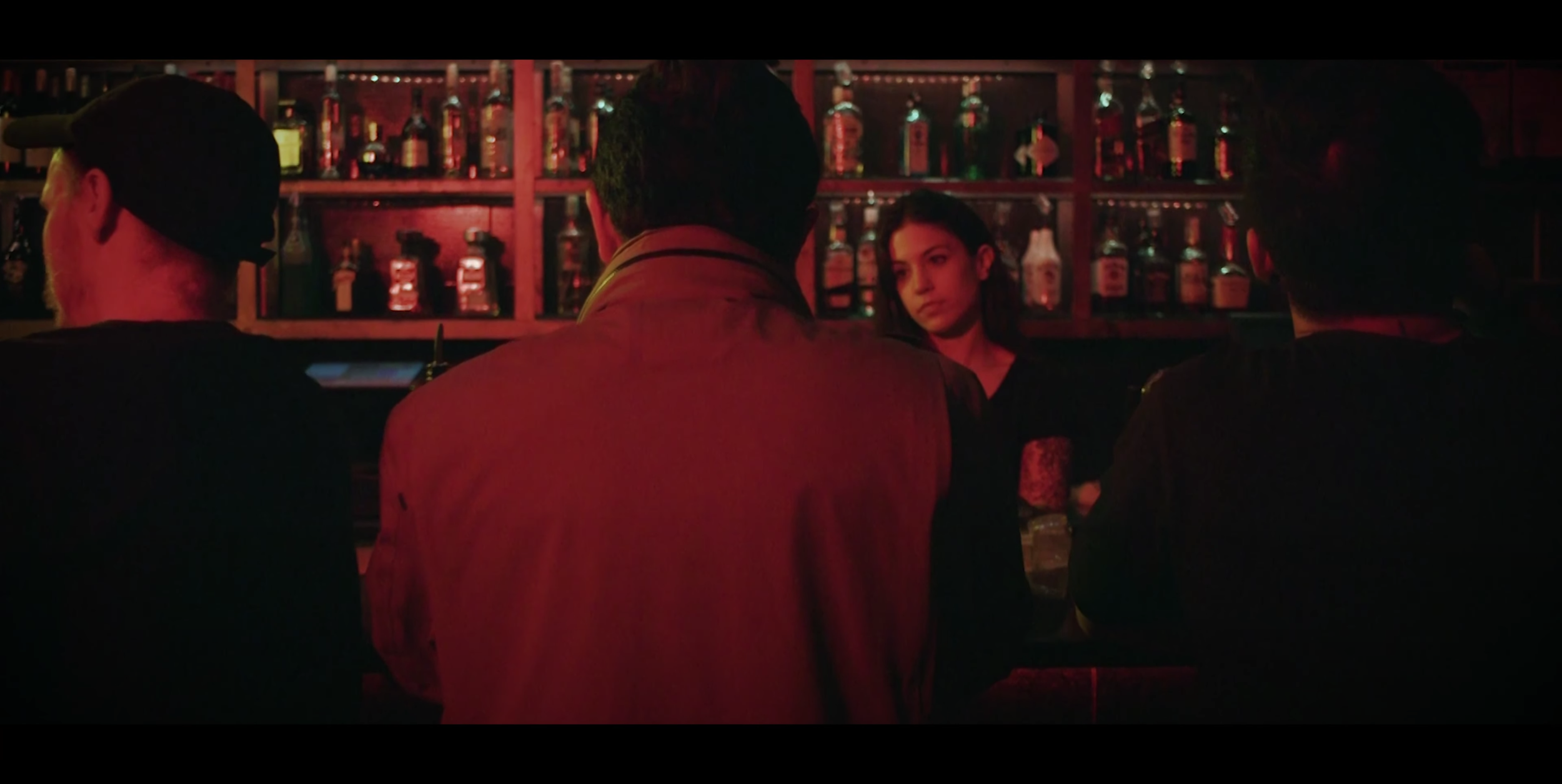 A bar scene with three men sitting at the counter and a woman standing behind the bar, all in low lighting with red tones, bottles of alcohol visible on shelves in the background.