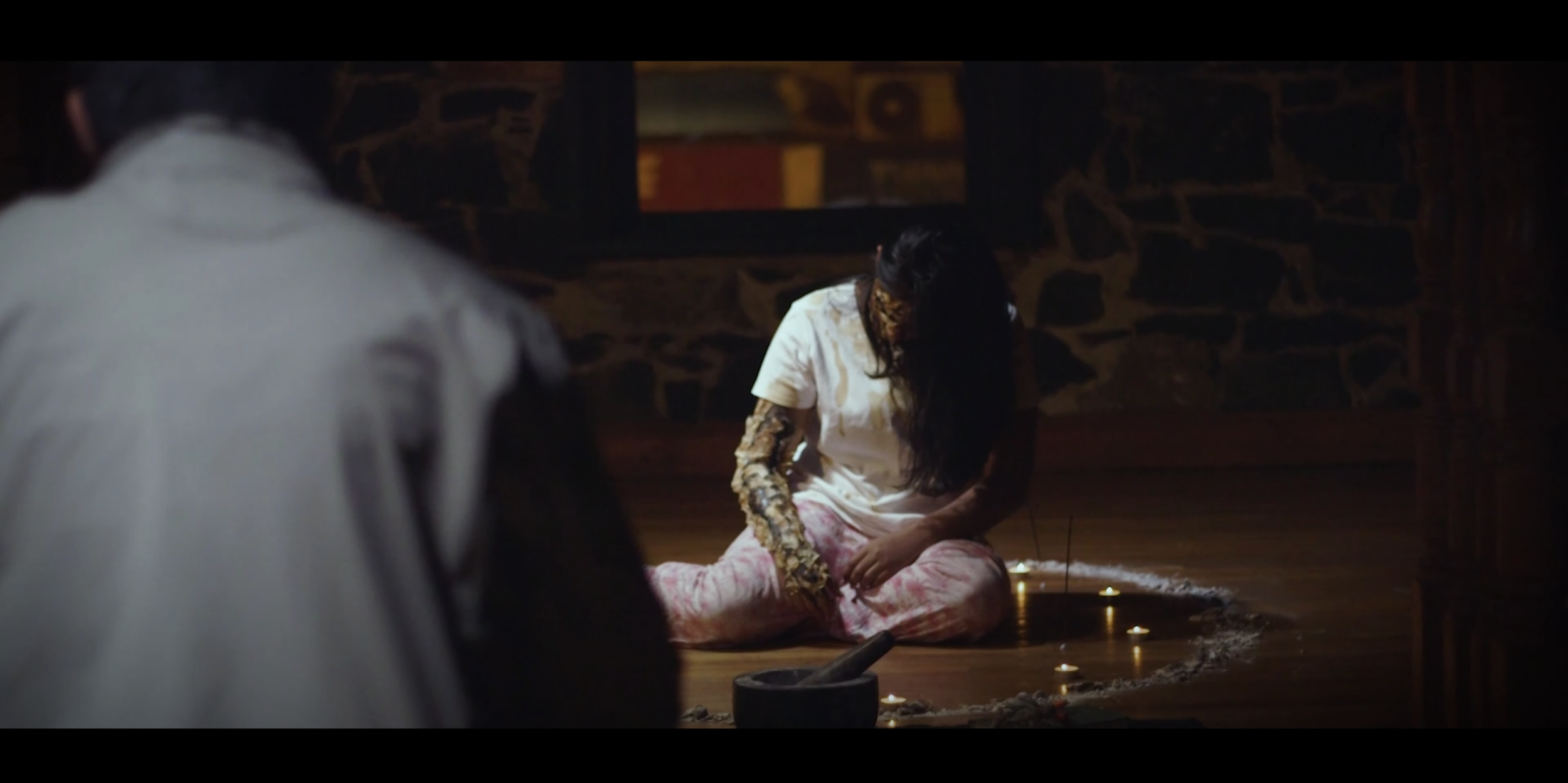 A woman with long dark hair, wearing a white shirt and pink pajama pants, is sitting on the floor with her head bowed and injuries or scars on her arms and face, surrounded by lit candles in a circle in a dimly lit room with stone walls. A person in a white shirt is partially visible in the foreground, facing her.