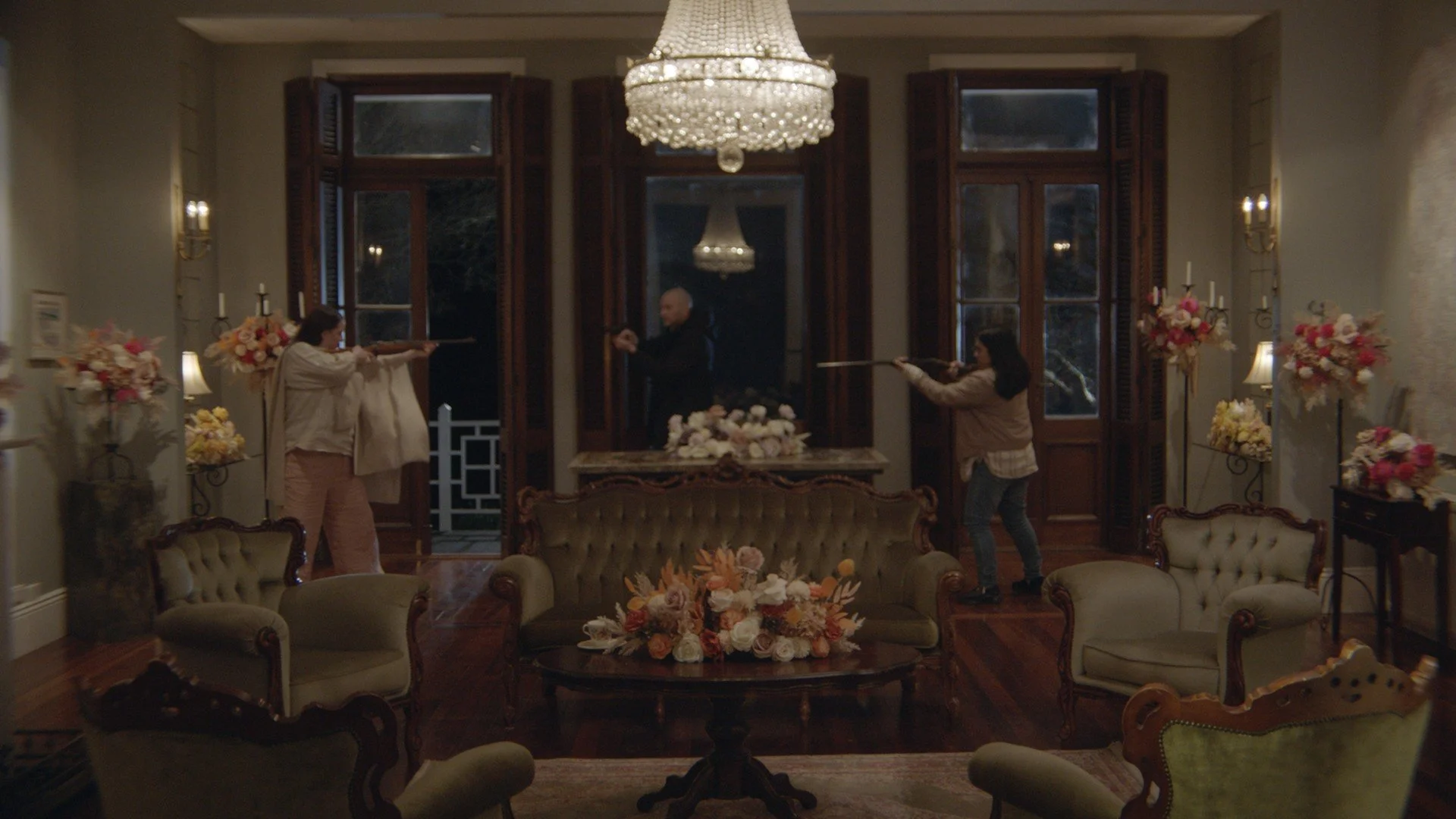 Three people inside a decorated living room aiming rifles at each other, with vintage furniture, floral arrangements, and chandeliers.