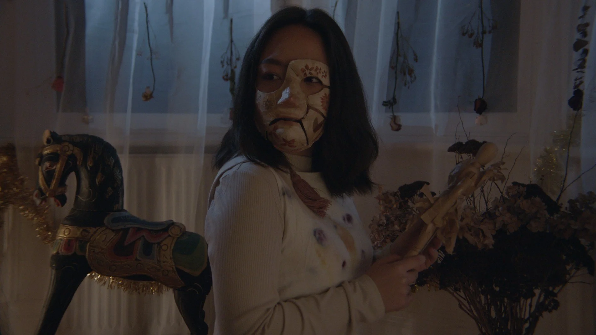 A woman wearing a decorative face mask and a cream-colored top, standing indoors with dried flowers and a vintage carousel horse.