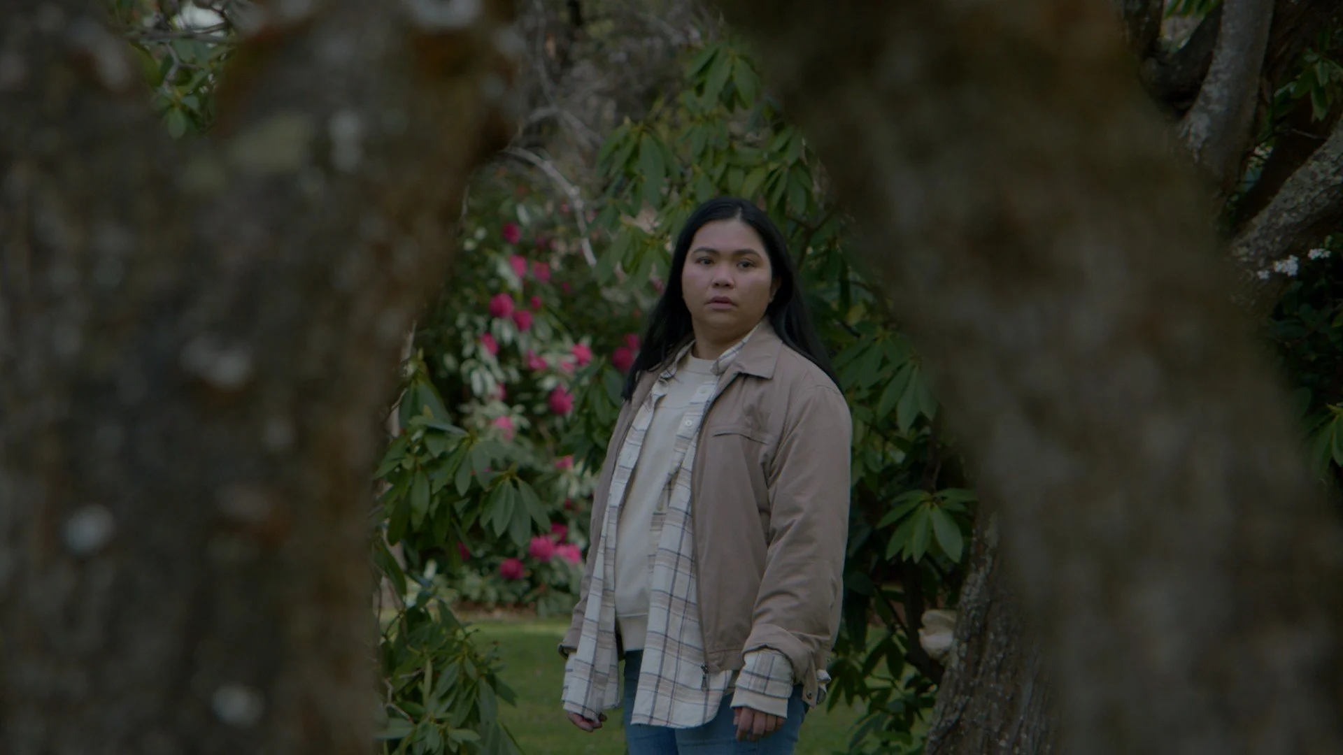 A woman with long black hair in a beige jacket and plaid shirt standing between two tree trunks in a garden with pink and white flowers.