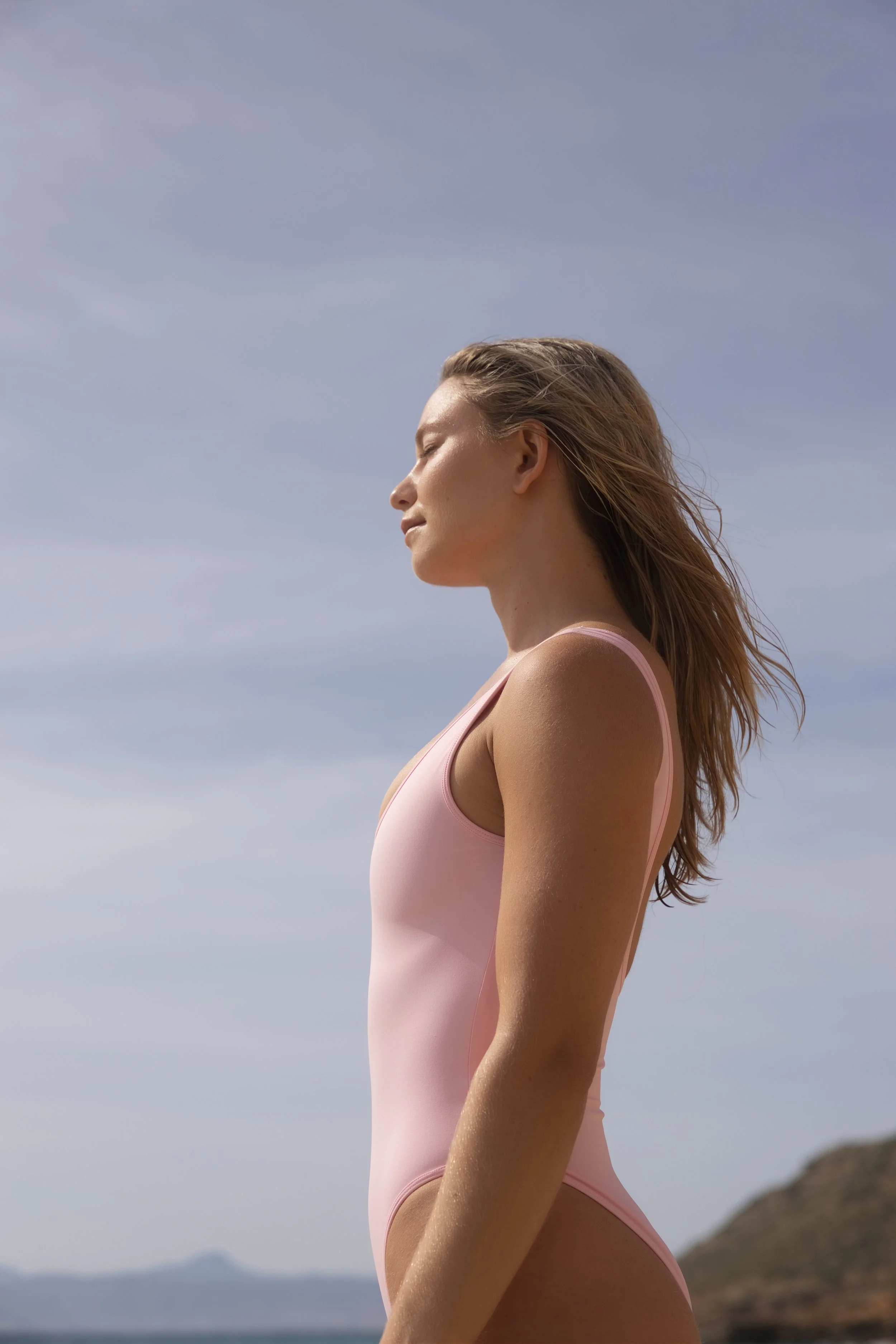 Side profile of a woman with closed eyes and long, wavy hair, wearing a light pink tank top, standing outdoors with a clear sky and distant mountains in the background.