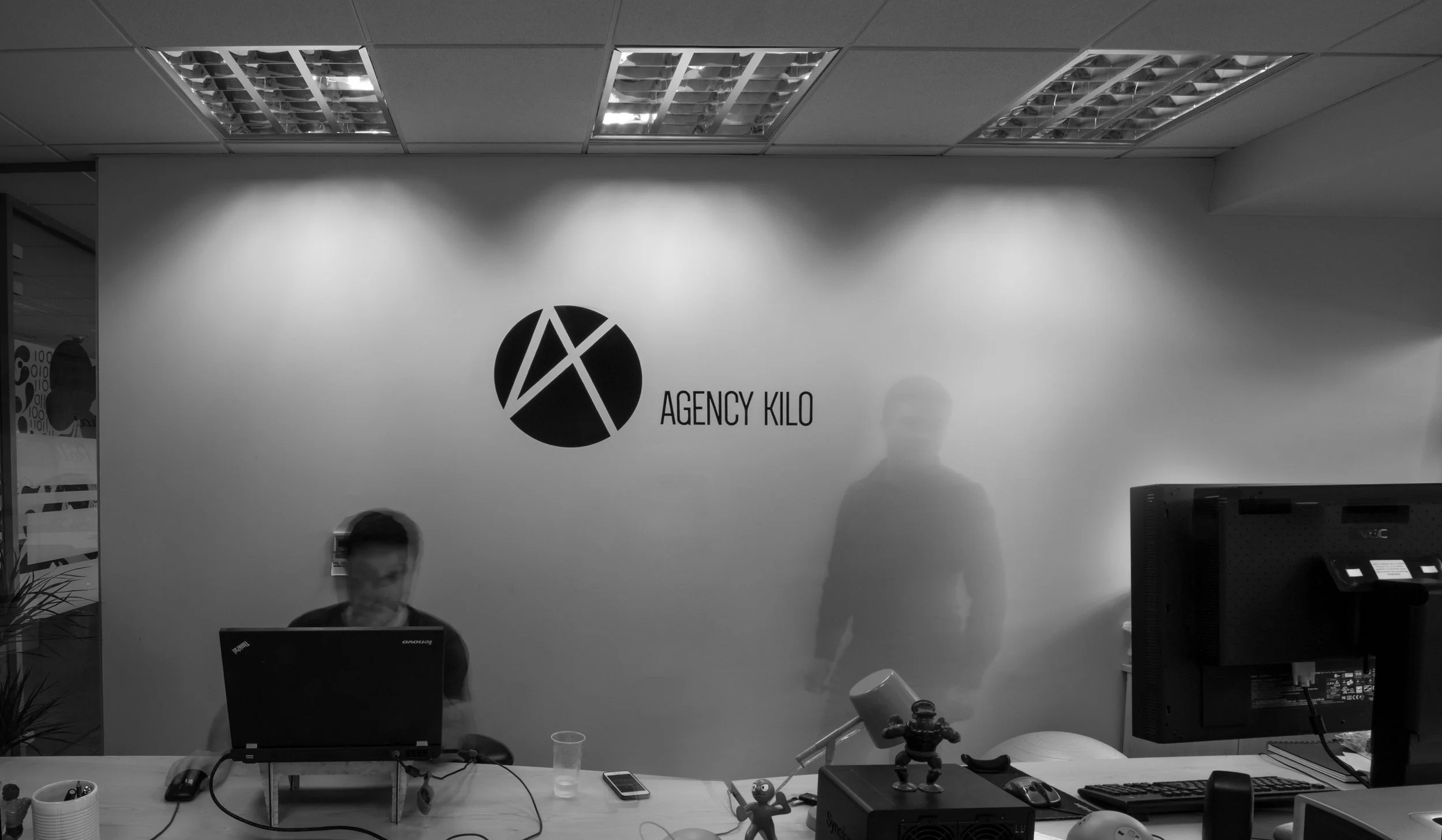 Office reception area with a frosted glass wall, two people, one sitting at a desk with a laptop and the other standing, logo reading 'Agency Kilo' on the wall, desk items including a computer monitor, keyboard, mouse, smartphone, and office decor.