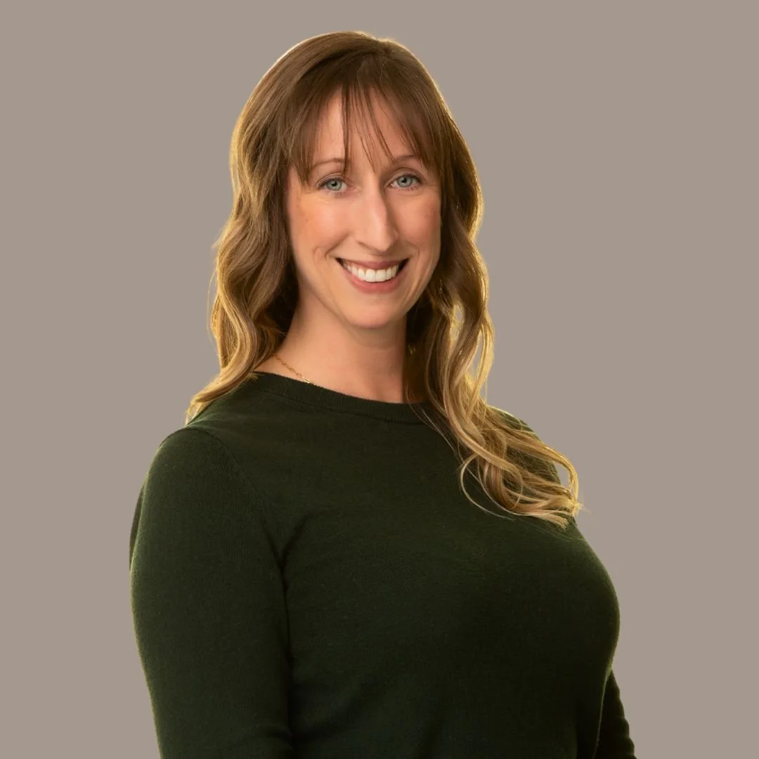 A woman with shoulder-length brown hair, blue eyes, and a friendly smile, wearing a navy blazer over a white blouse, standing against a plain neutral background.