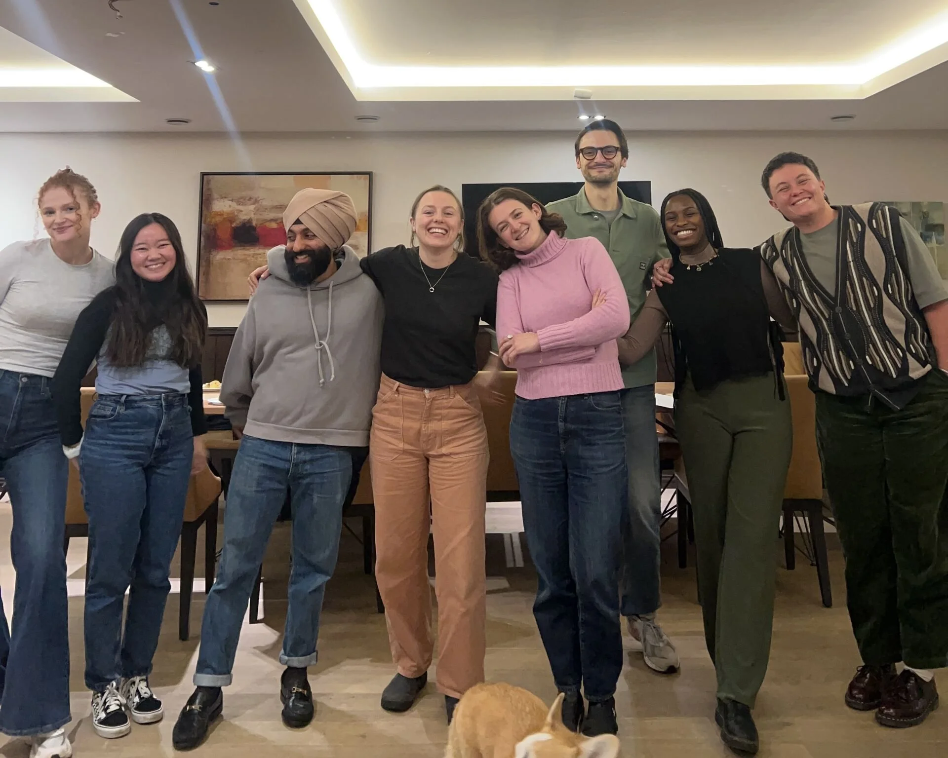 From left to right, a tall red haired white woman; a short, black haired asian person; short man in a turban; a white woman with brown hair; white woman with auburn hair; tall white man with glasses; tall black woman; tall white person.