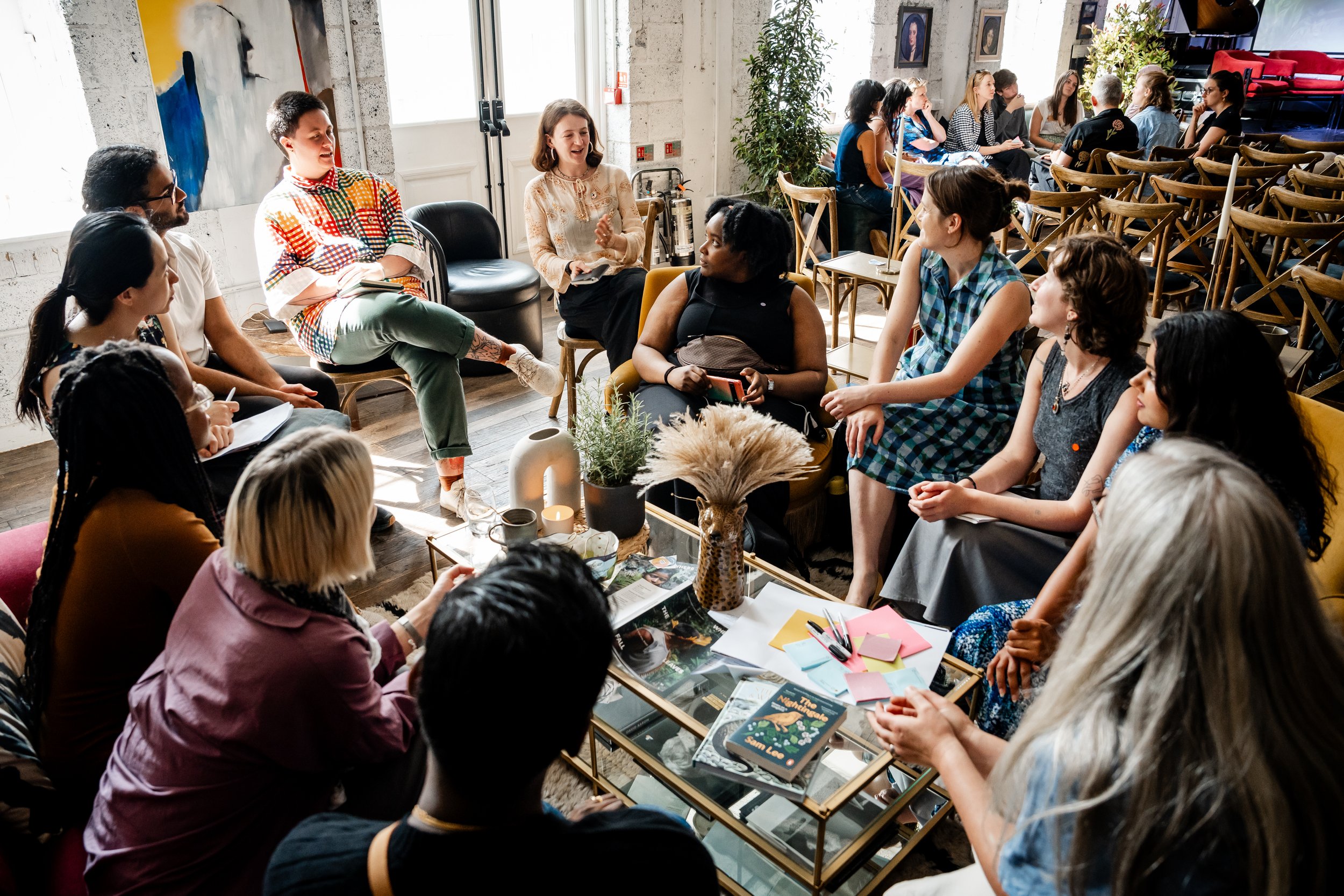 12 people with diverse skin tones, hair colours and styles sat in a circle deep in conversation. In the middle of a the circle is a table covered in magazines and stickers.