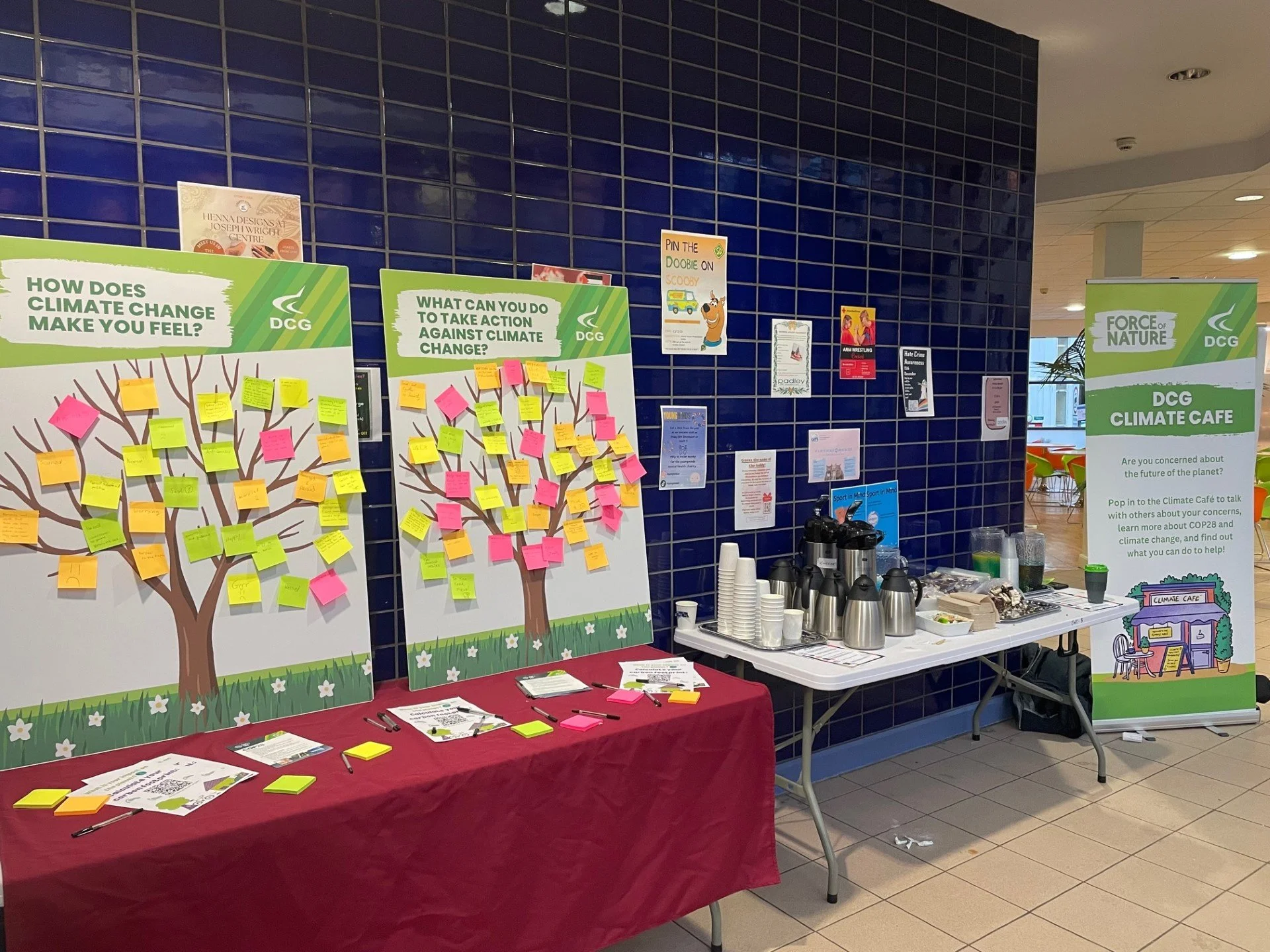 Two upright boards on a table say "How does climate change make you feel?" and "What can you do to take action against climate change?". There are sticky notes with people's responses. On the right is a table with refreshments.