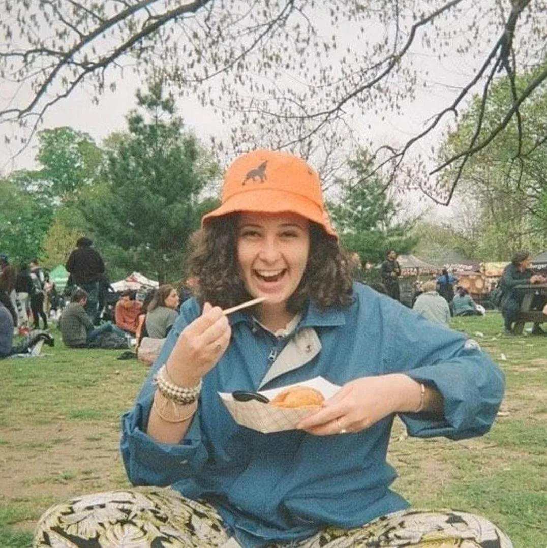 A woman with curly hair wearing an orange bucket hat is smiling at the camera whilst holding a plate of food.
