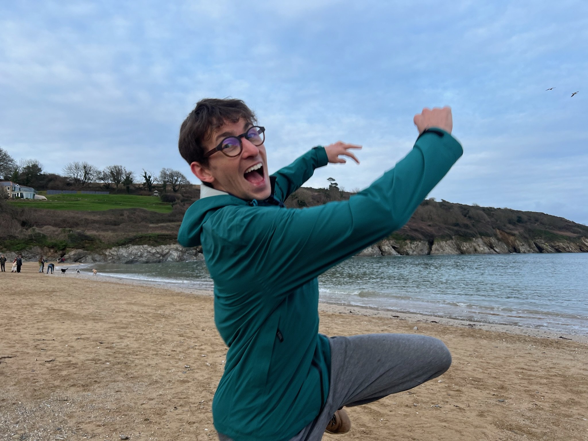 A man with short dark hair and round glasses is jumping and smiling on a beach.