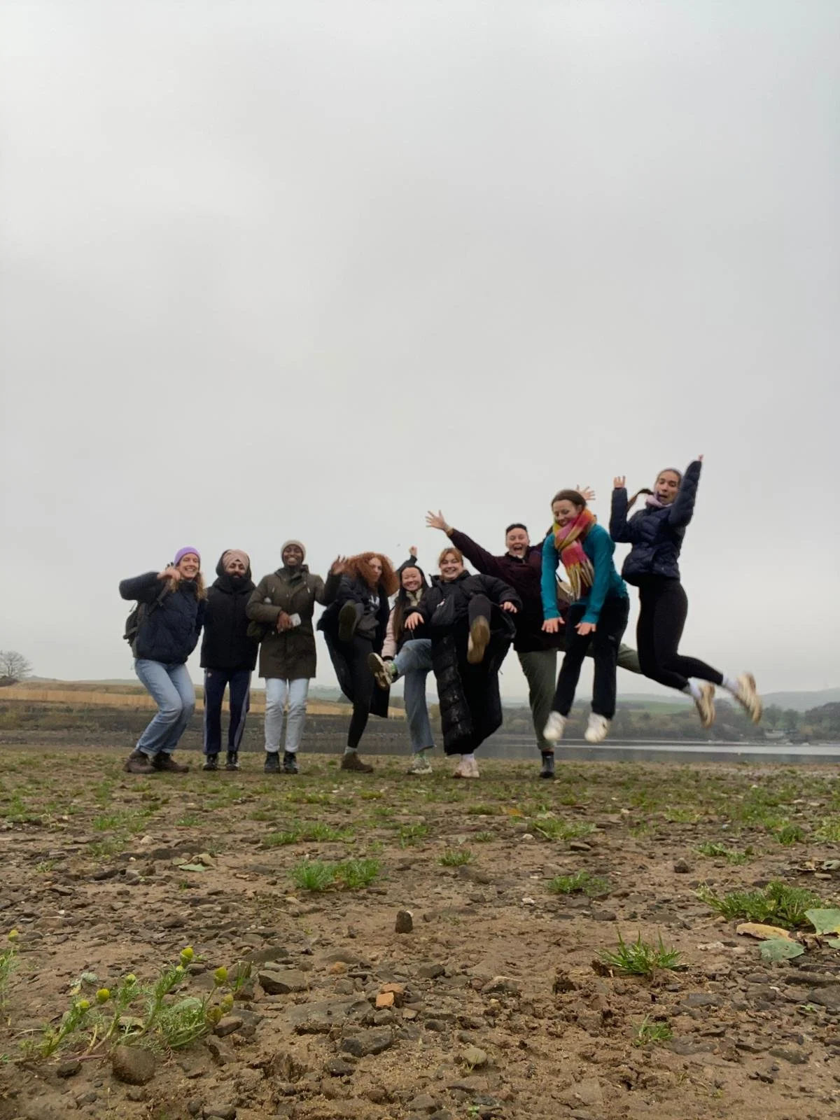 9 people jumping in the air for a team photo in front of a lake. The ski is grey and people are wearing scarves and hats.