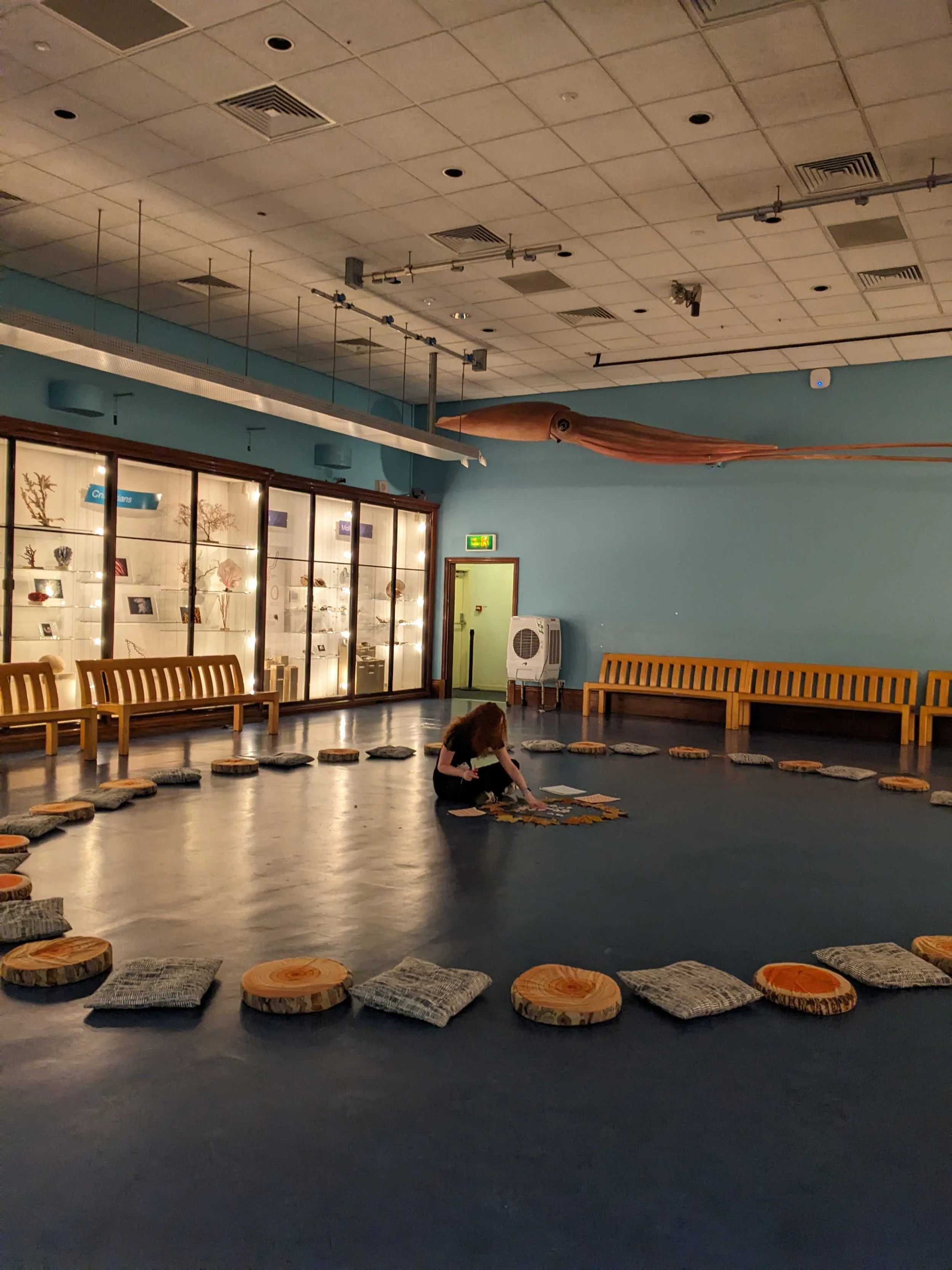 Red haired woman sat crossed legged in the middle of a circle of cushions inside a room that is painted blue. At the top of a room, hangs a model of a giant squid.