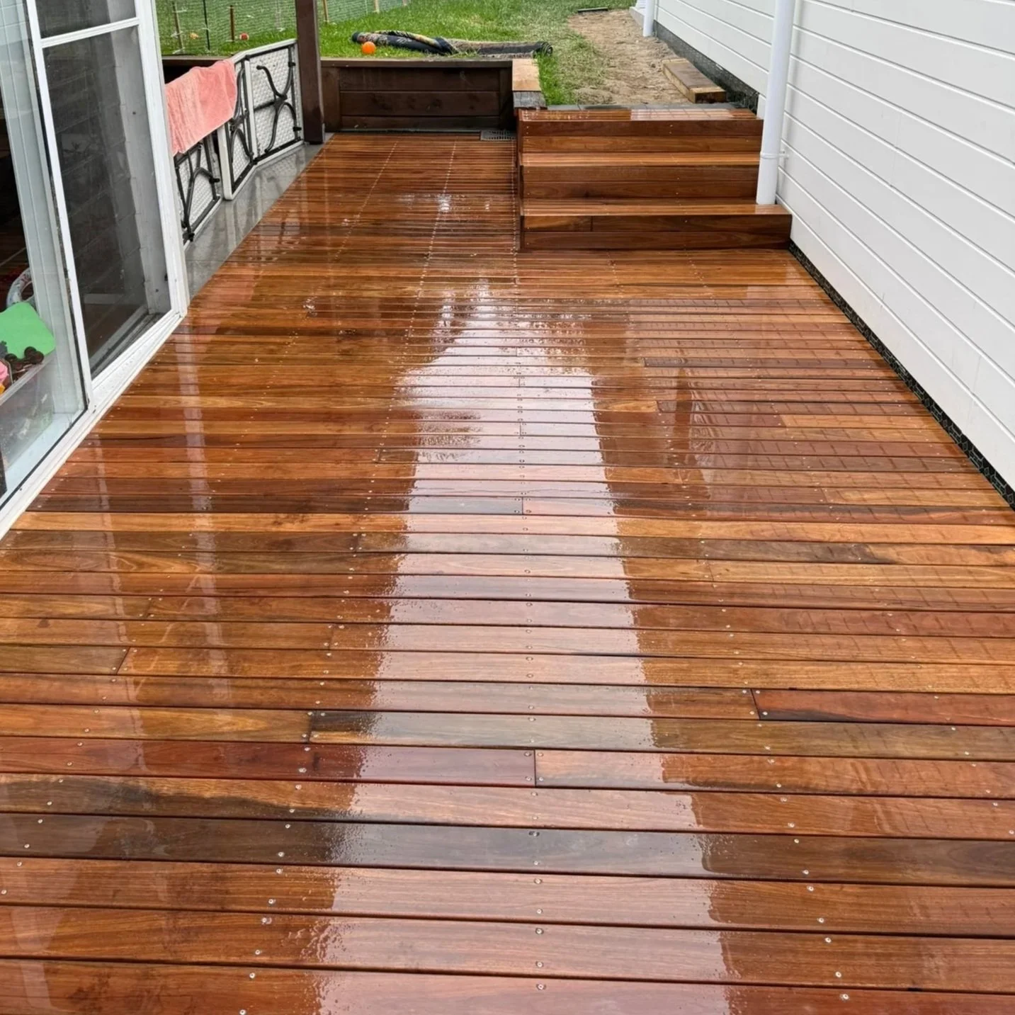 Rain-soaked wooden deck outside a house, with reflections of the sky on the wet surface, and a sliding glass door on the left.