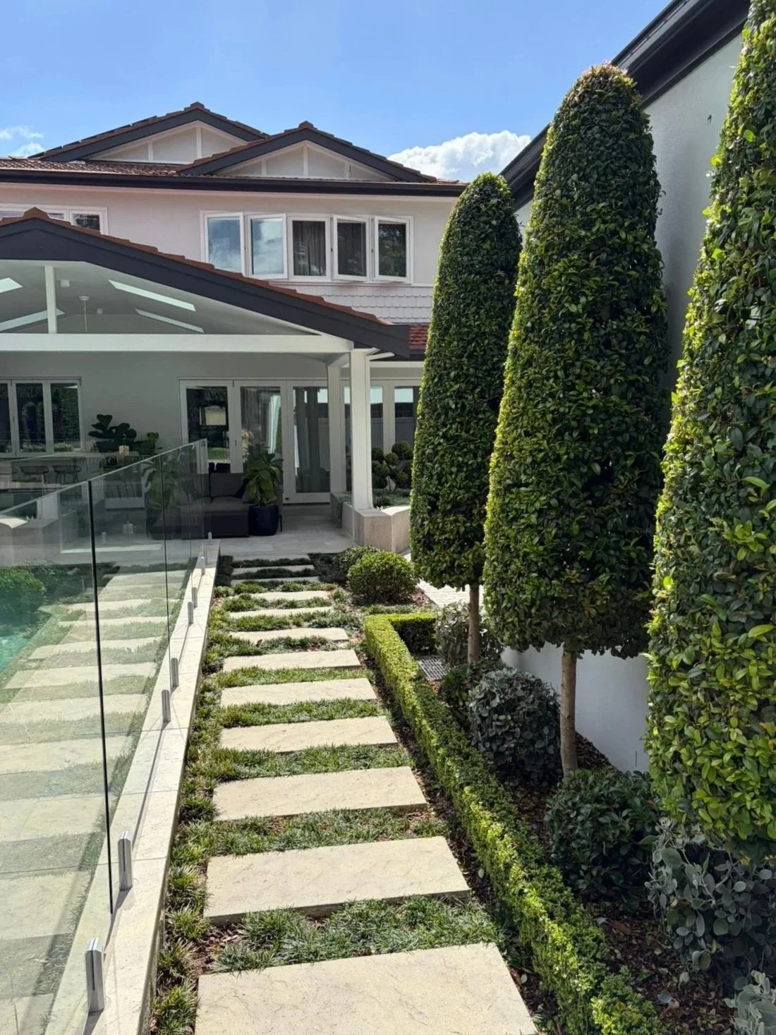 Modern backyard with a stone pathway, trimmed bushes, and tall, conical trees alongside a white house with a glass patio area.