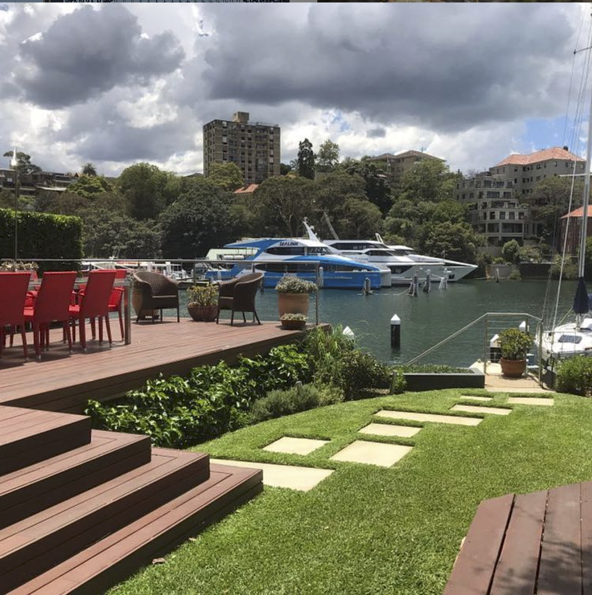 A marina with luxury yachts docked in the water, seen from a backyard patio area with red chairs, a wooden deck, and stepping stones on a grassy lawn, under a cloudy sky.