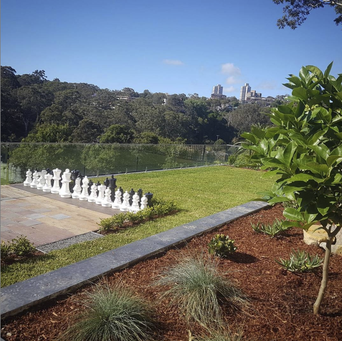 A garden with a large chess set, green grass, plants with reddish mulch, and a backdrop of trees on a hillside with tall buildings in the distance under a blue sky.