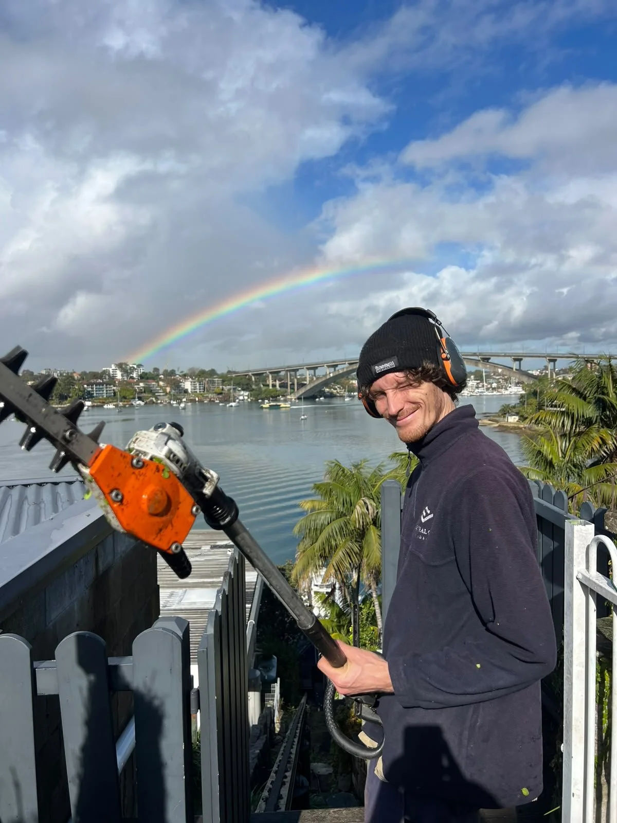 A man in a black hat and headphones using cutting equipment outdoors near water, with a rainbow, clouds, a bridge, boats, palm trees, and city buildings in the background.