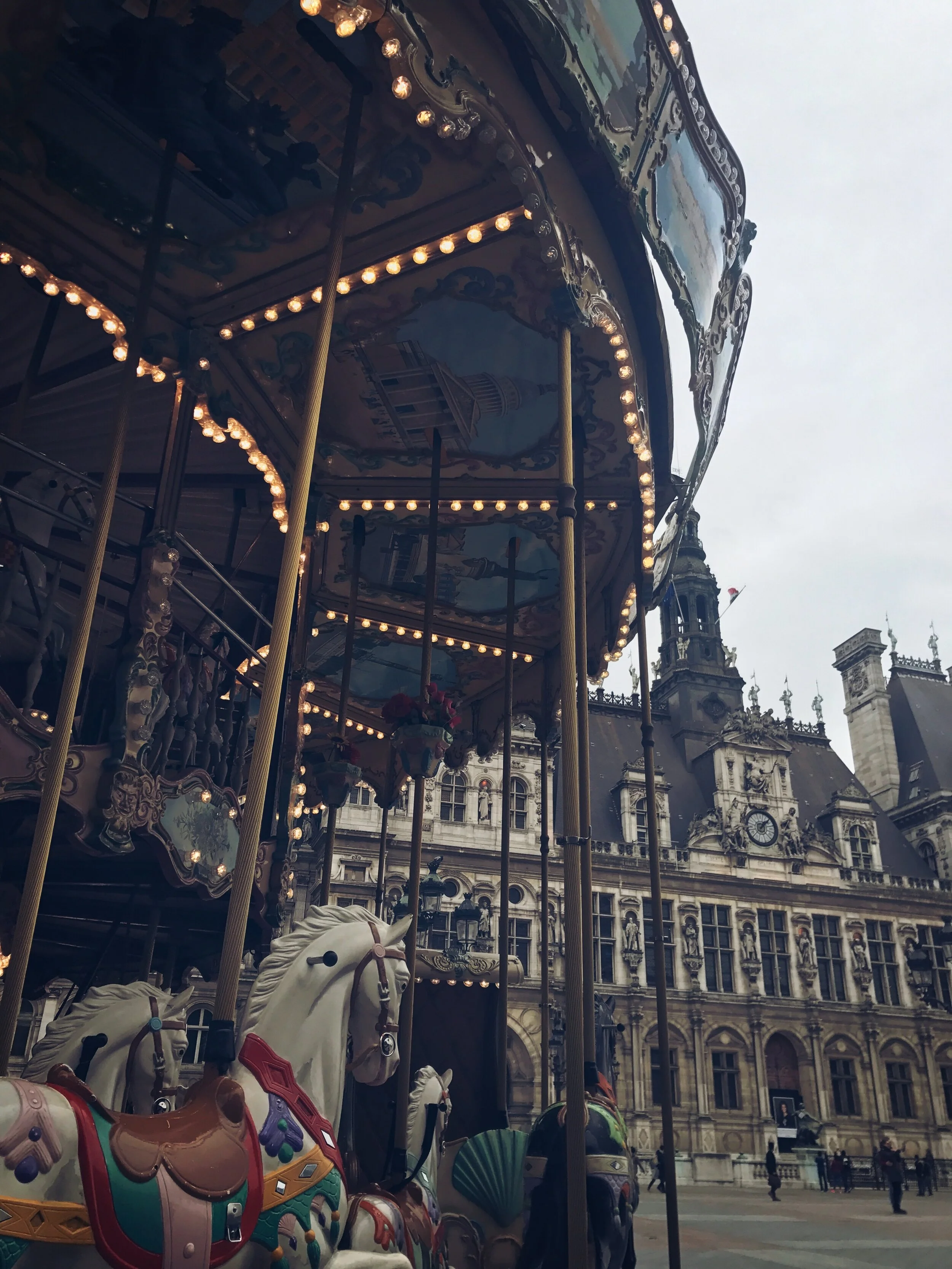A vintage carousel with white horses and decorative lights in front of a historic European building. Paris