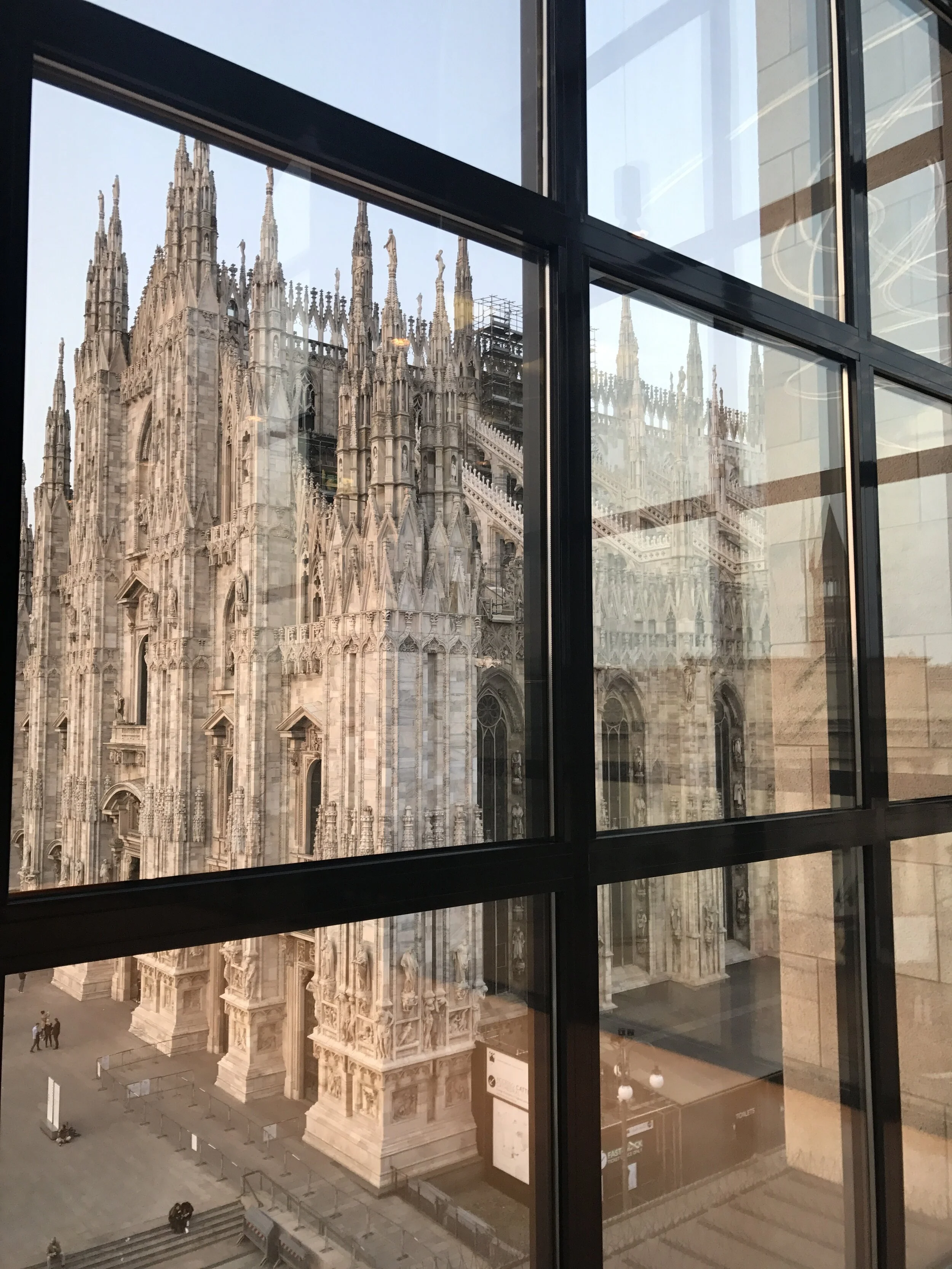 View of Milan Cathedral through glass window showing ornate Gothic architecture with spires and detailed sculptures.