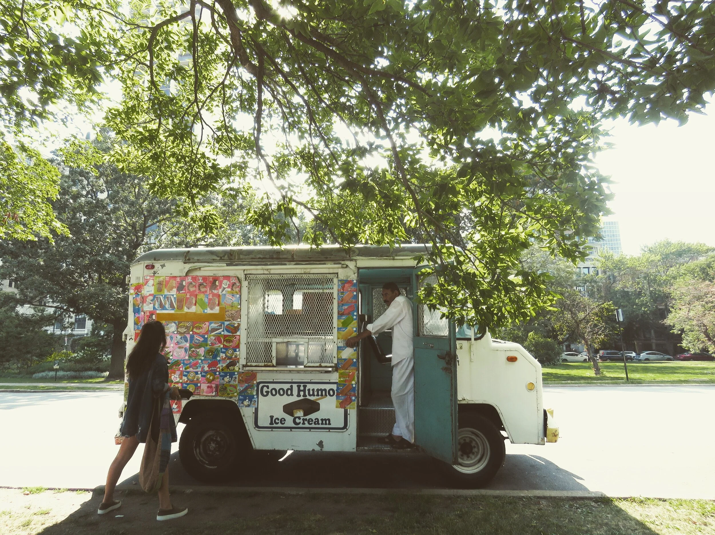 An ice cream truck parked on a street under a large tree. A man in white is serving ice cream from the truck, and a woman with long hair and a black coat is standing nearby, waiting. The truck has colorful decorations and a sign that reads 'Good Humo