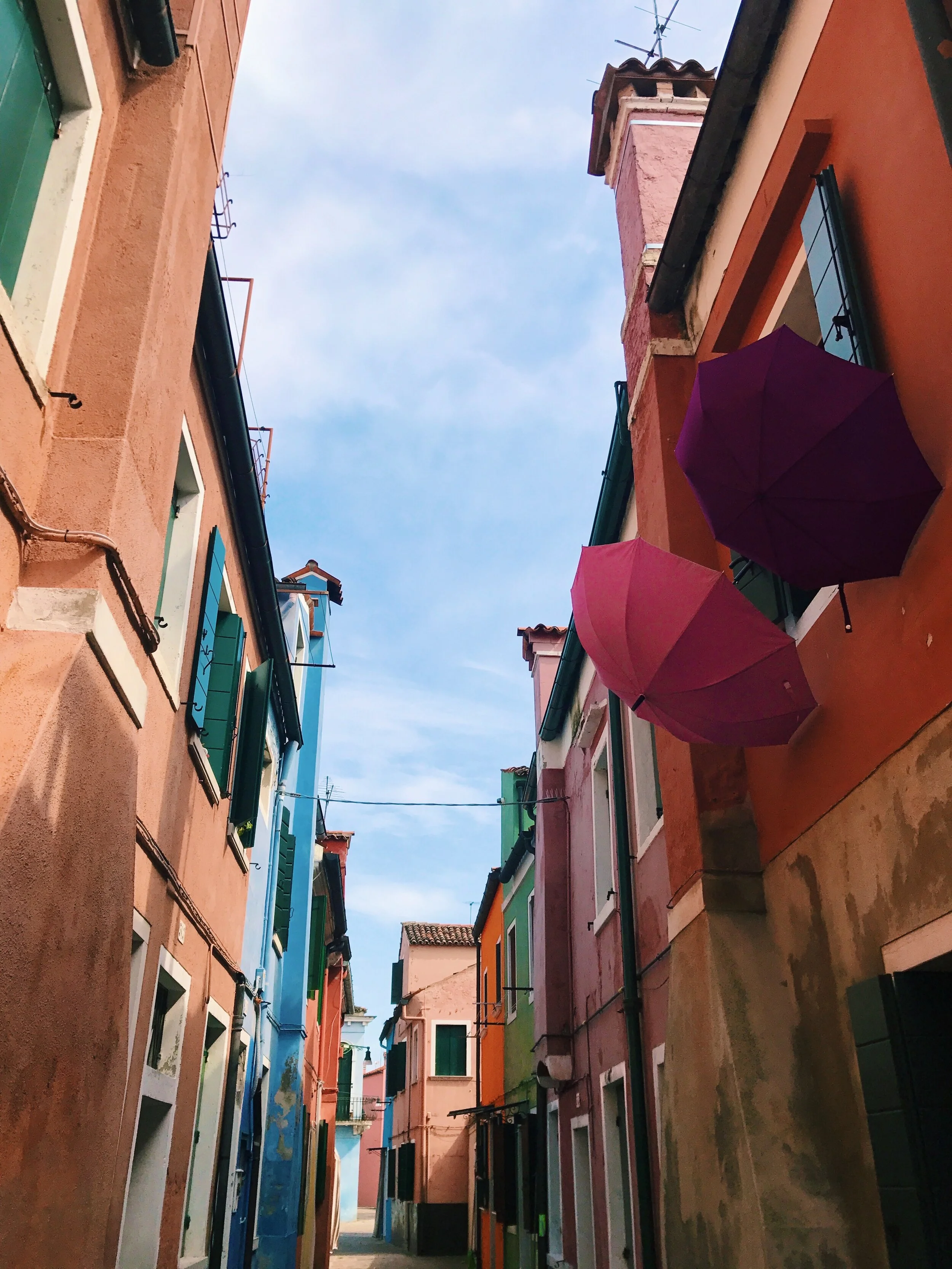 Colorful buildings with green shutters along a narrow street, with two purple umbrellas attached to the building on the right and a pink umbrella on the left. Venice Moreno