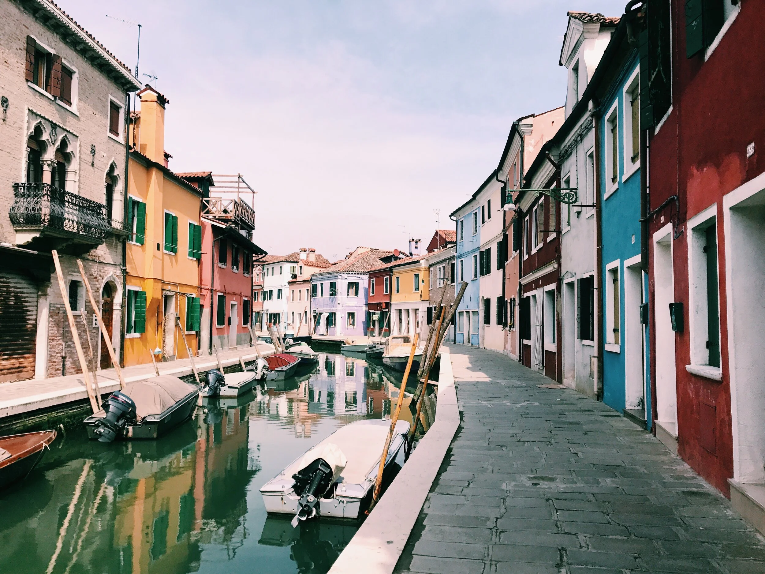Colorful buildings lining a small canal with boats docked along the water and a cobblestone pedestrian pathway on the right side. Venice islands