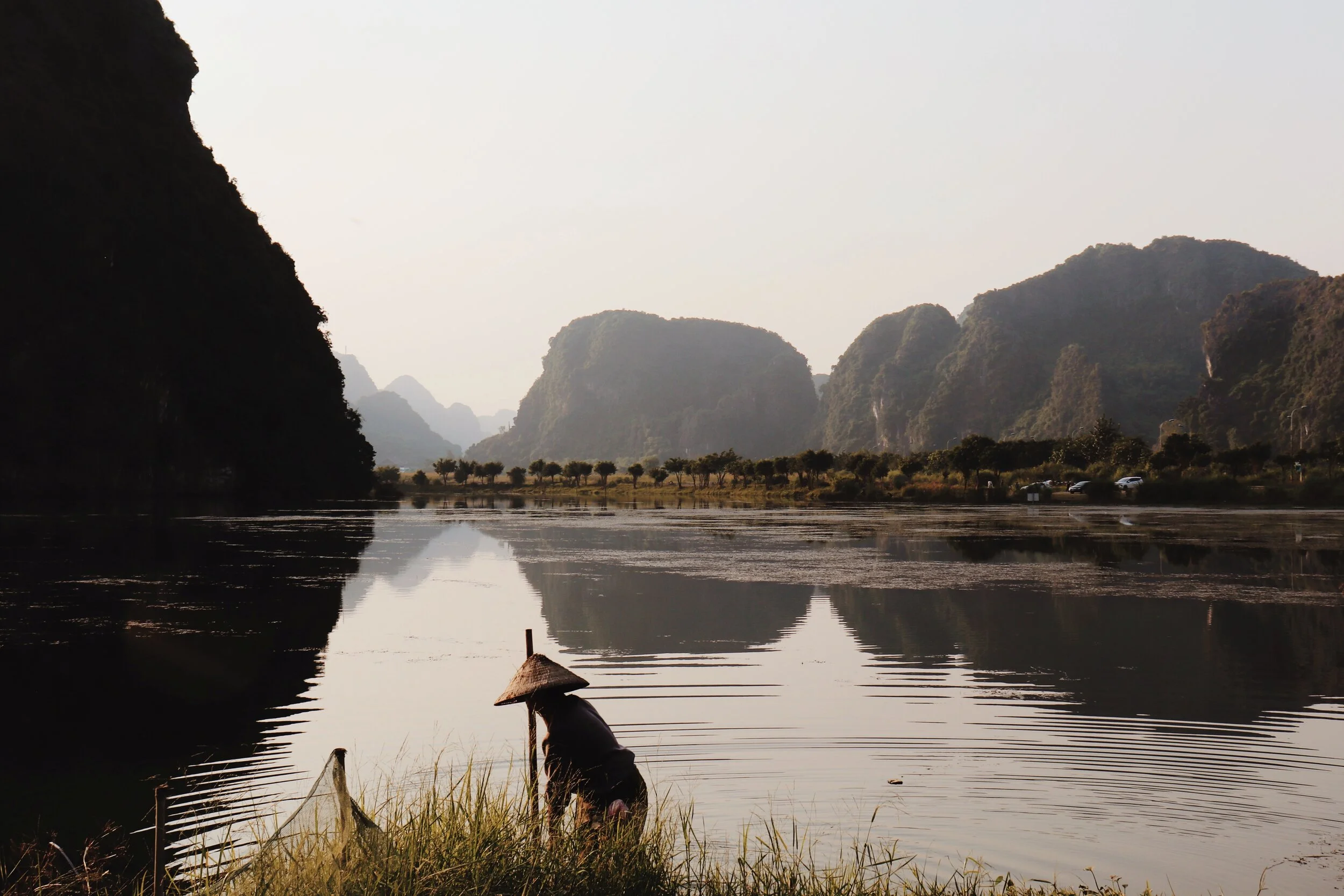 Person with a conical hat leaning over a river, surrounded by mountains and trees in a peaceful landscape. farmer in Vietnam