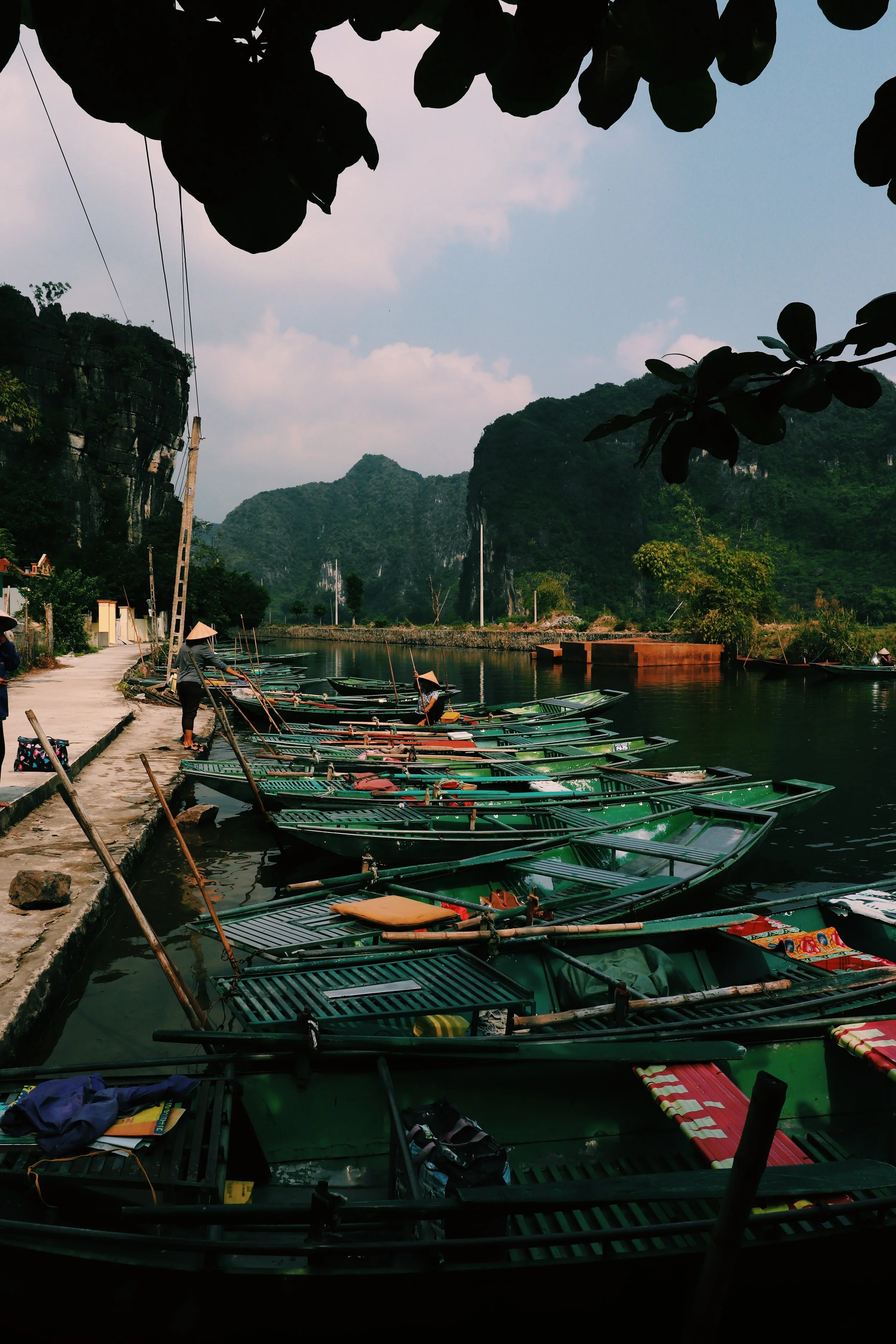 A waterfront scene with multiple green boats docked along a paved pathway, surrounded by lush green mountains and trees, with a cloudy sky overhead. Vietnam