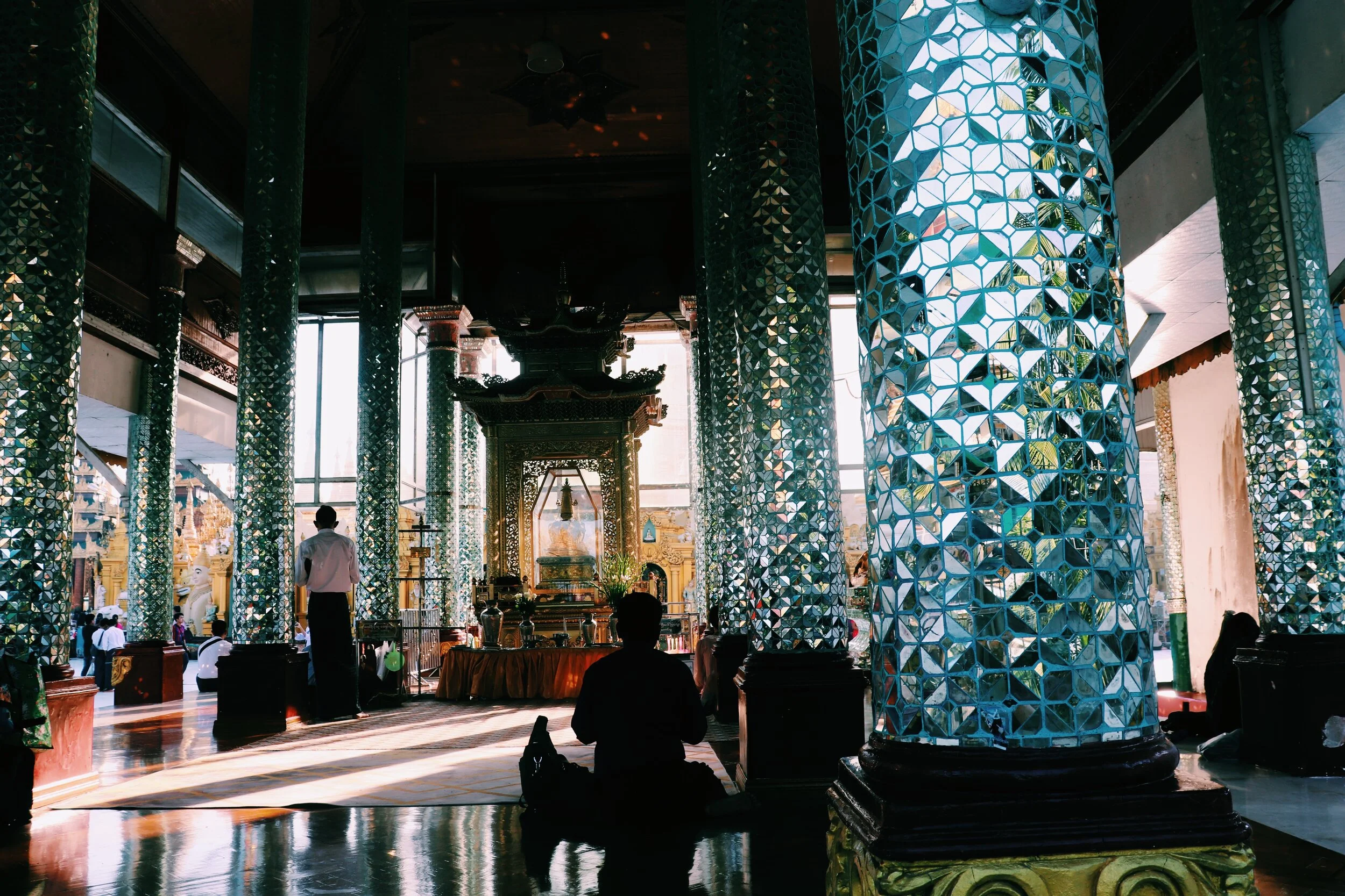 A person sitting cross-legged on the floor inside a temple with ornate columns made of reflective mosaic tiles, an altar at the center, and a person standing nearby in the background. Myanmar Yangon