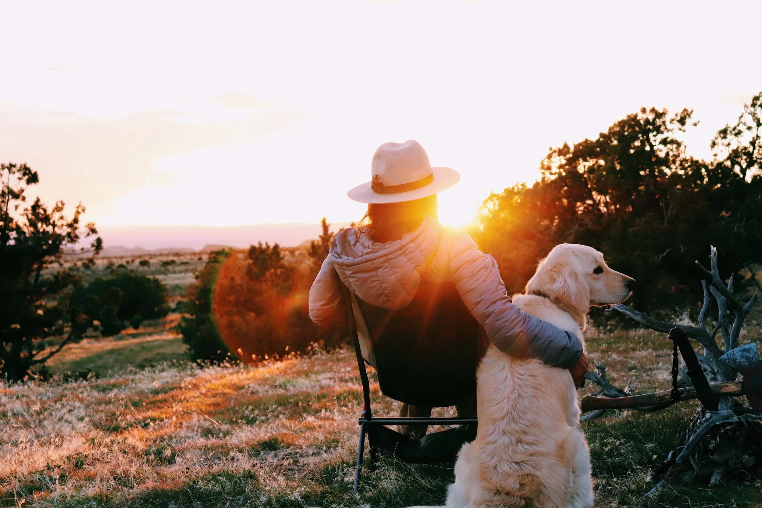 A person in a jacket and hat sitting on a chair outdoors at sunset with a golden retriever dog, surrounded by trees and open landscape. Utah camping