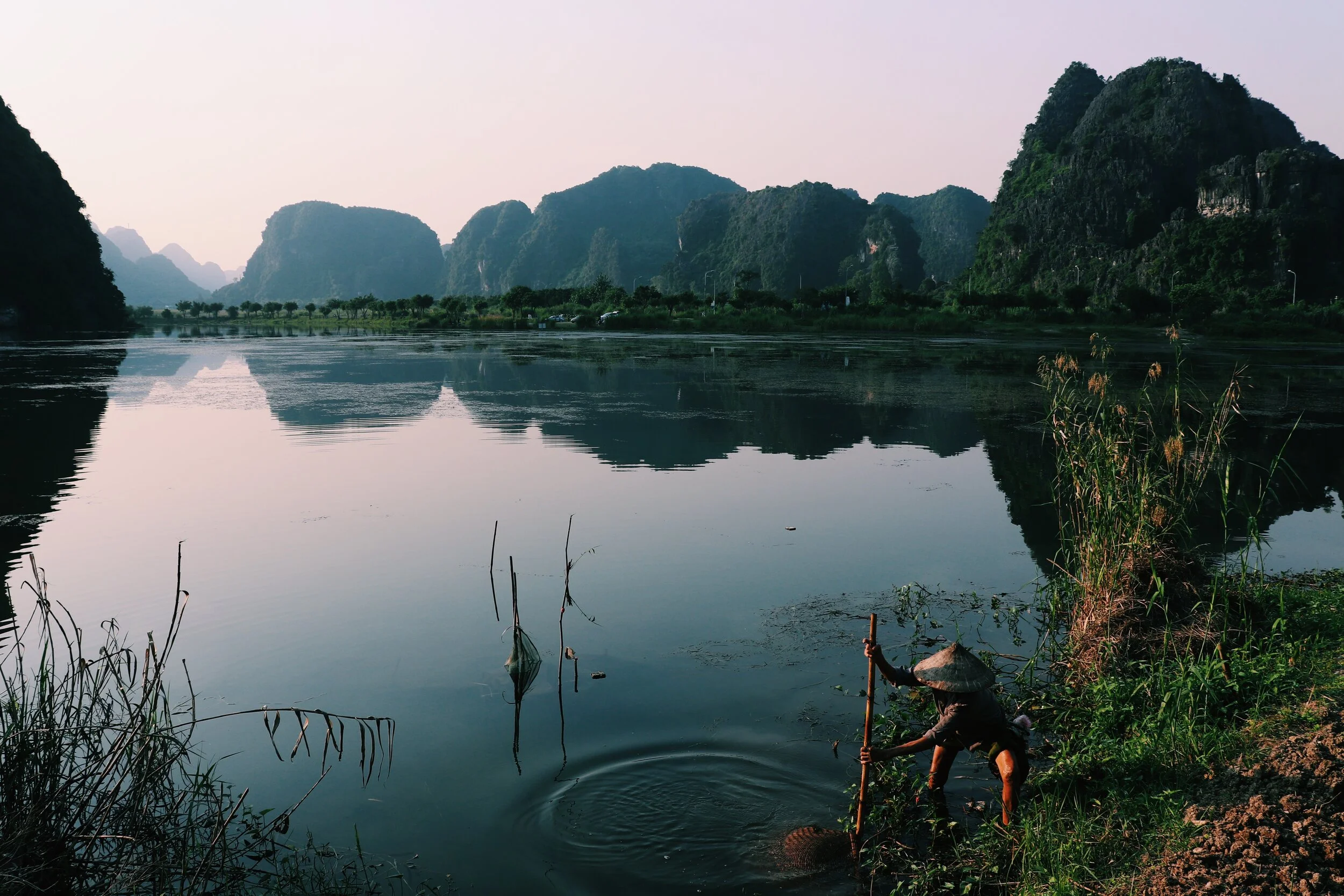 A person with a conical hat fishing by a river with mountains in the background. farmer in Vietnam 