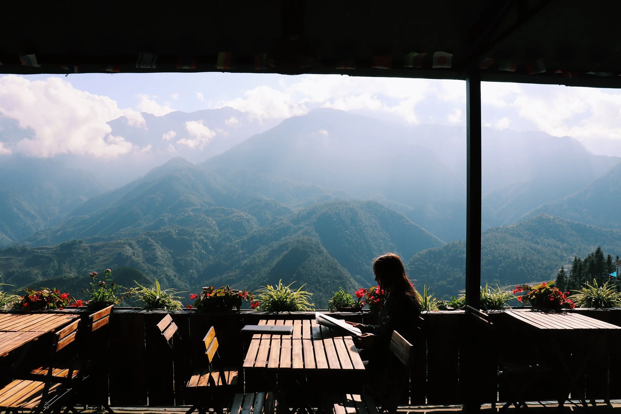 A person reading a book or menu at a wooden table on a terrace with a view of green mountains and clouds. Sapa Vietnam