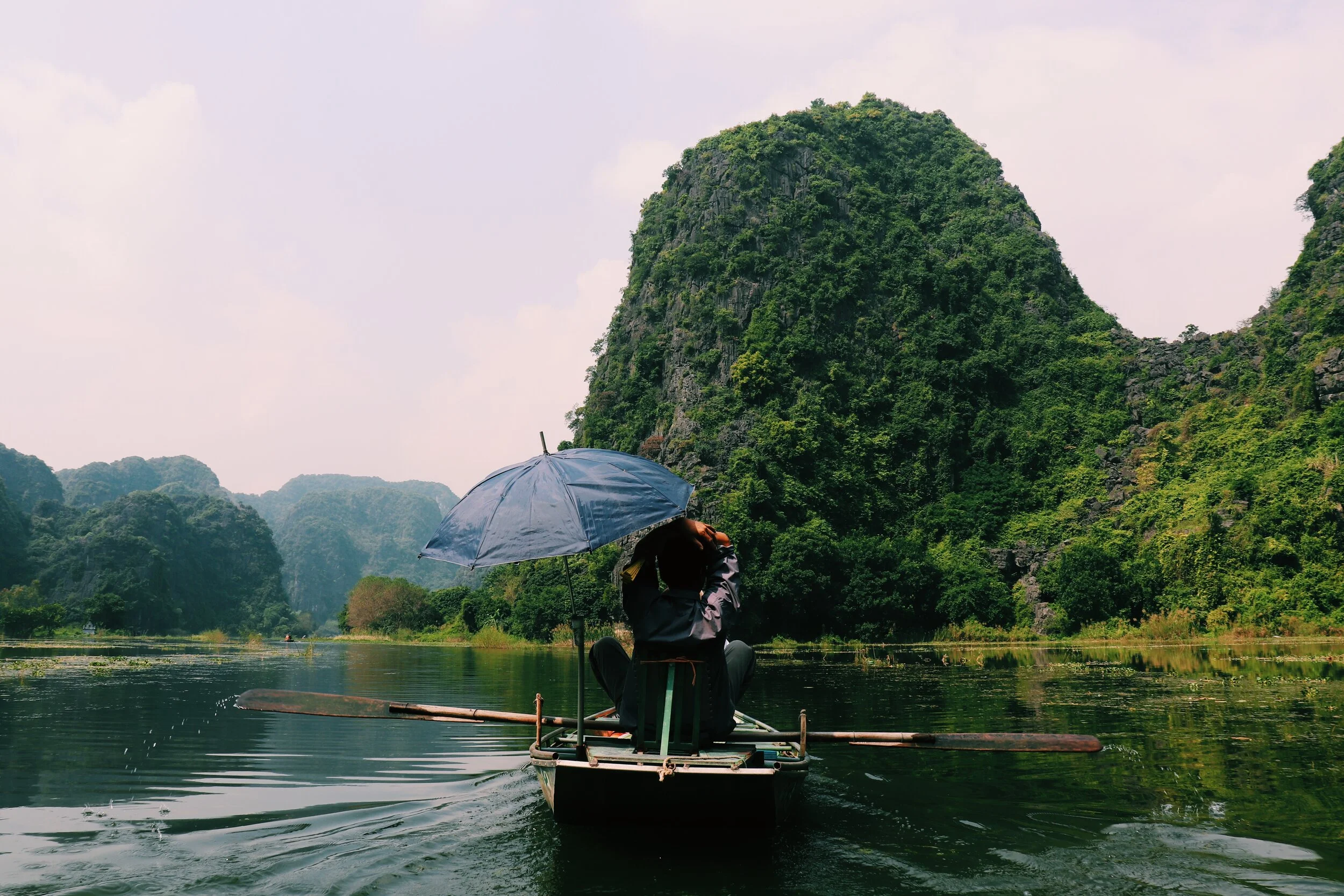A person sitting on a raft with a blue umbrella, rowing on a river surrounded by lush green mountains. Vietnam