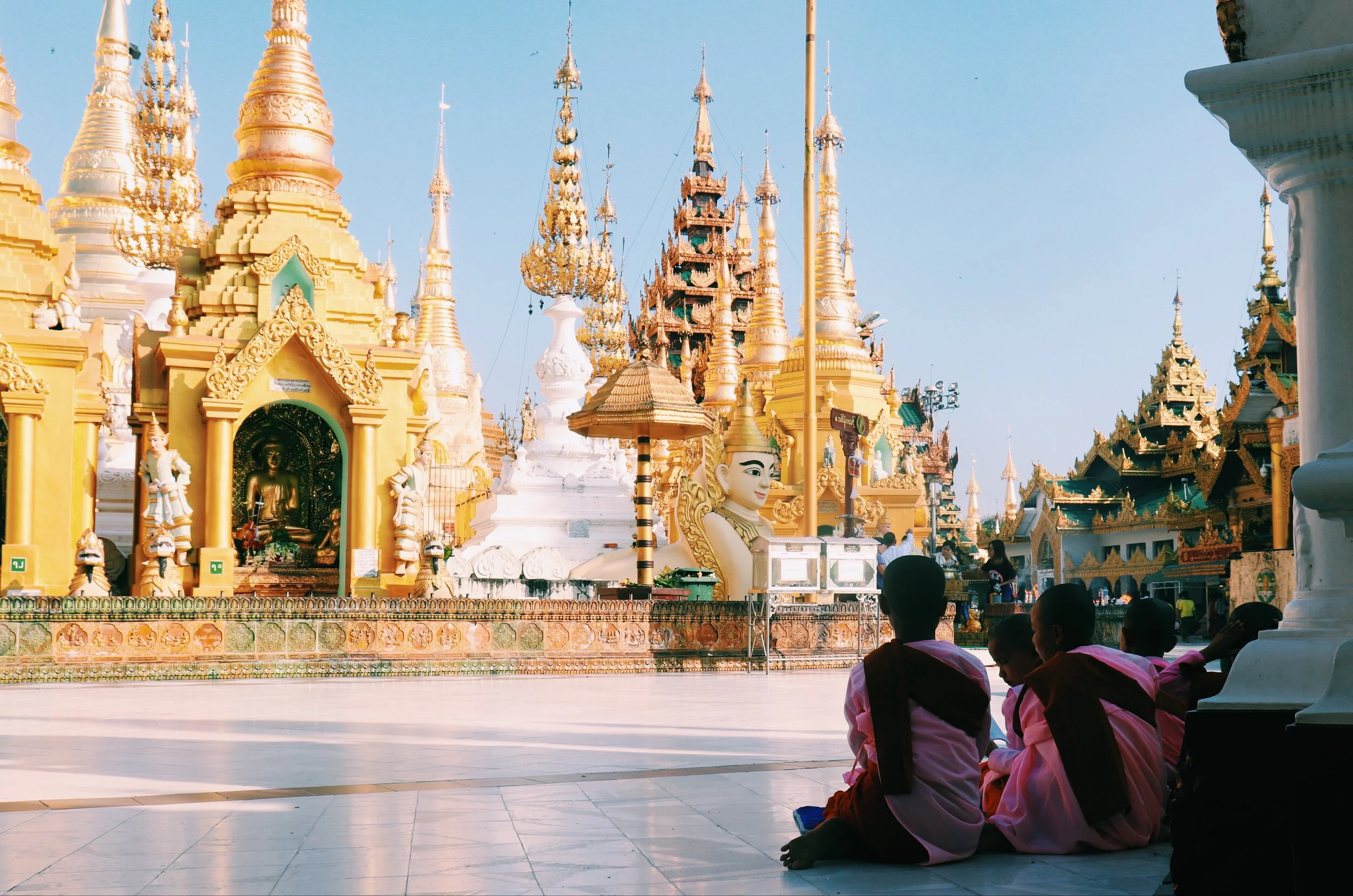 Group of children dressed in traditional Myanmar clothing sitting on the floor of a temple courtyard, with ornate pagodas and a large statue in the background. Yangon Myanmar 