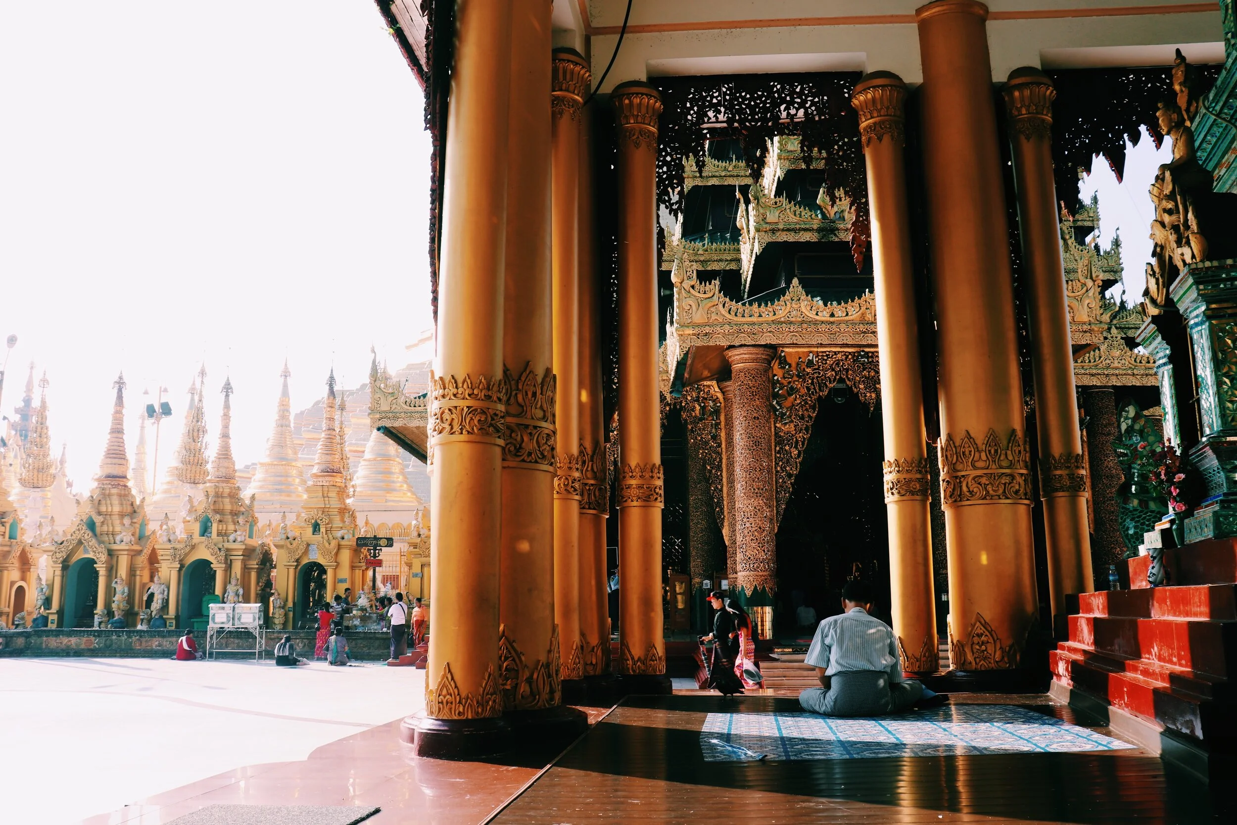 People praying inside a Burmese temple with gold columns and ornate decorations, with many white chedis or stupas in the background. Yangon Myanmar