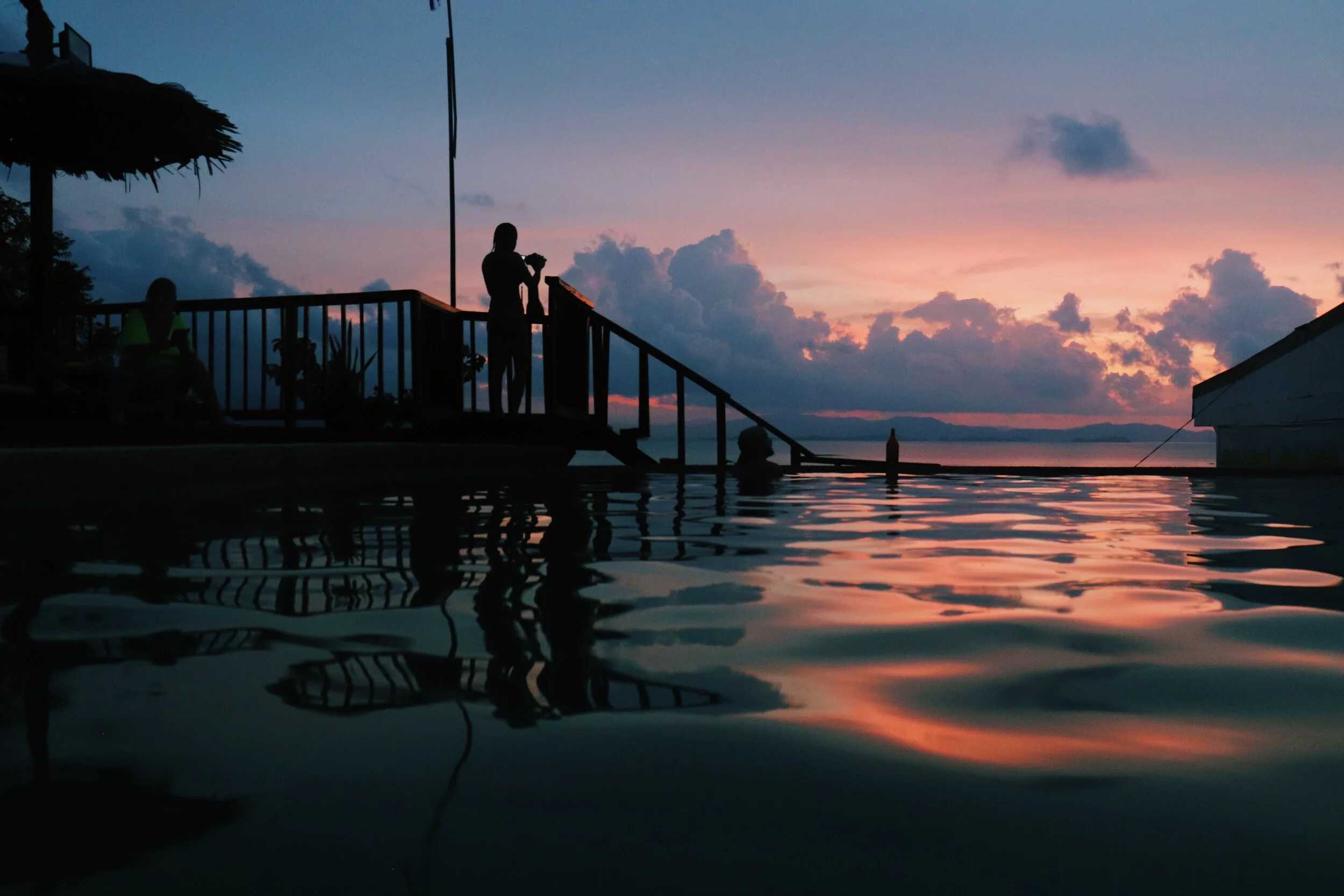 Silhouettes of people on a deck near water at sunset, with colorful sky and clouds reflected in the water. Philippines El Nido Palawan 